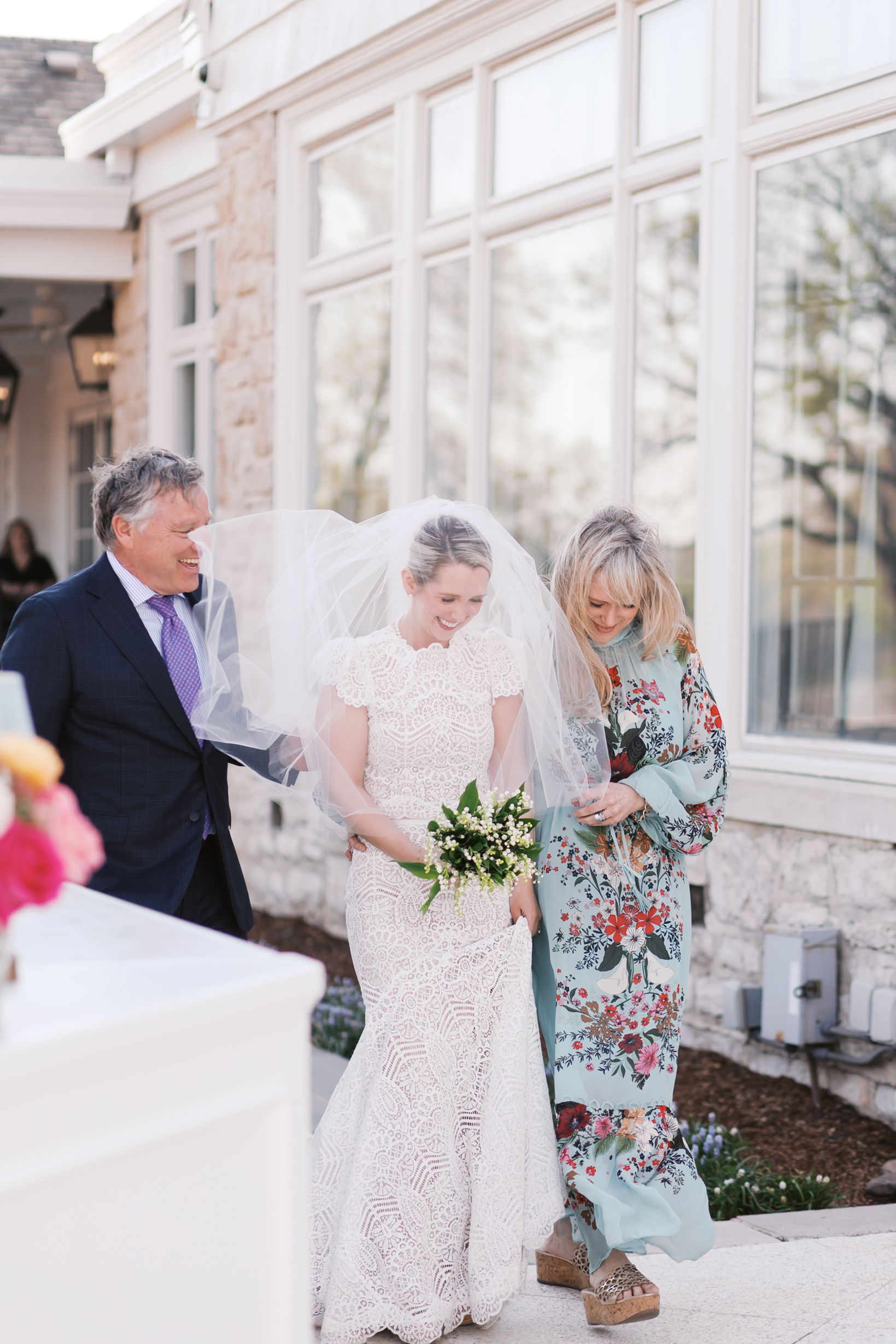 Bride walking outdoors with two adults holding her veil, wearing lace wedding dress and holding bouquet