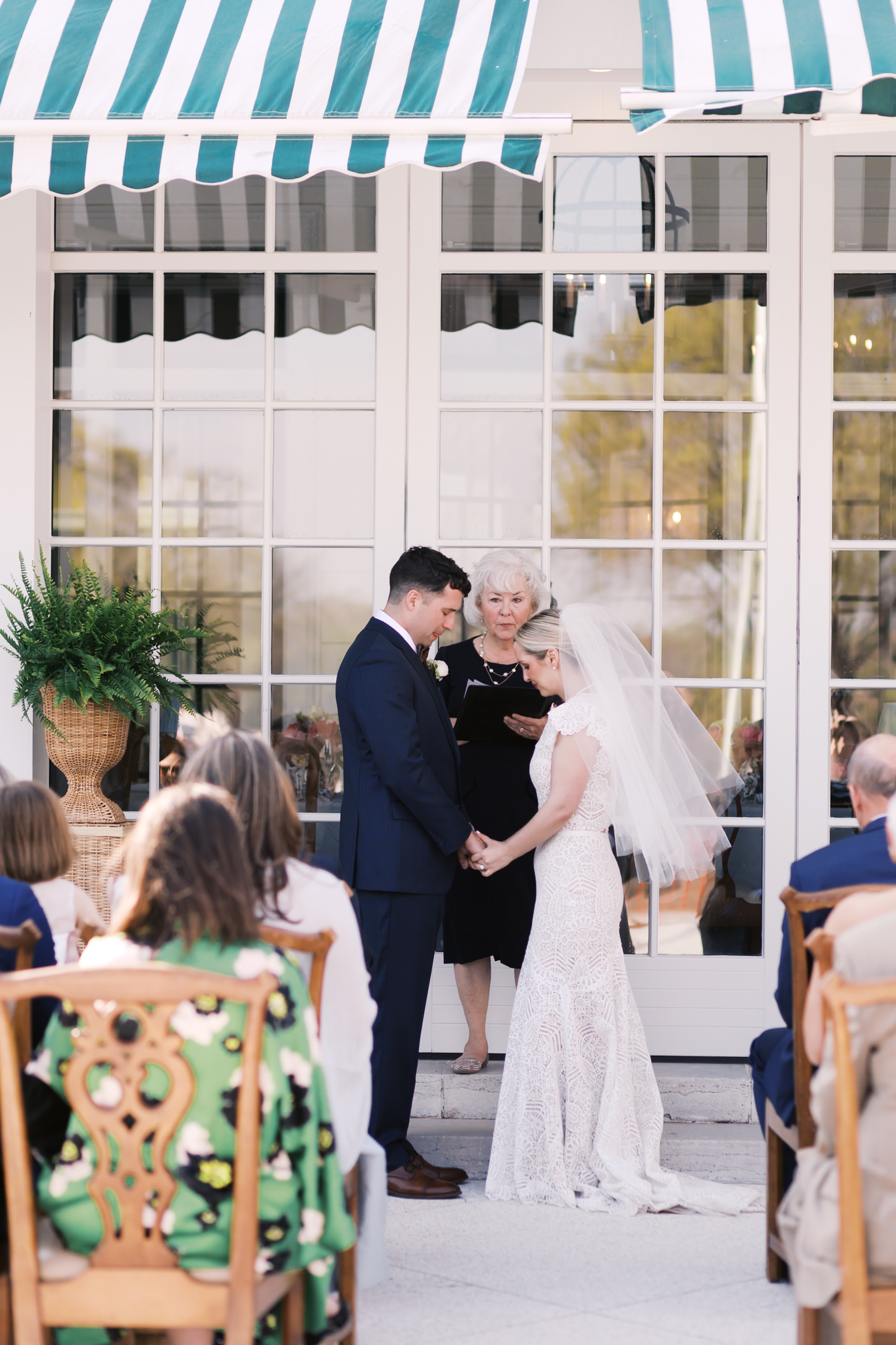 Bride and groom standing together during ceremony in front of glass doors and striped awning