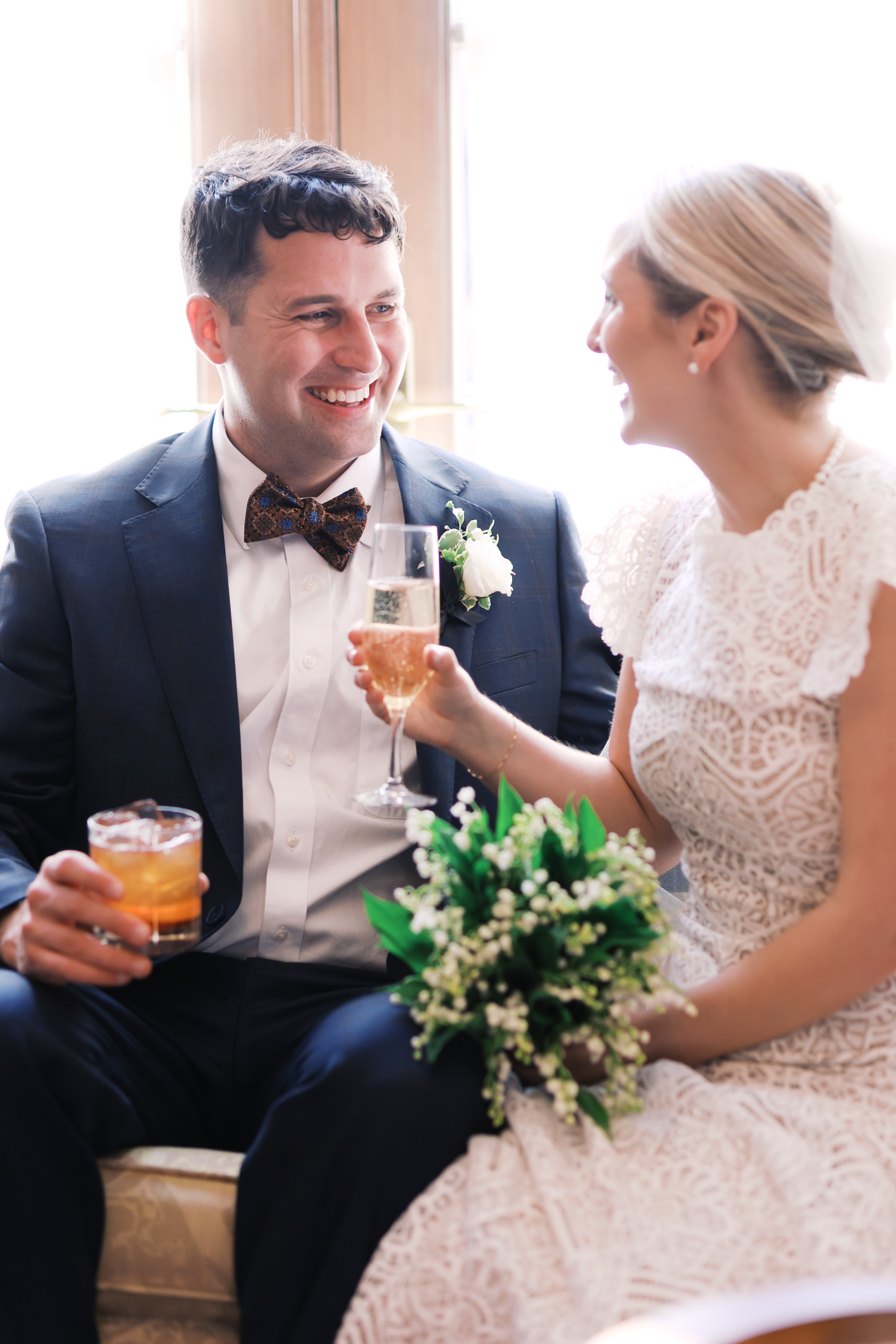 Bride and groom seated indoors holding drinks and smiling at each other
