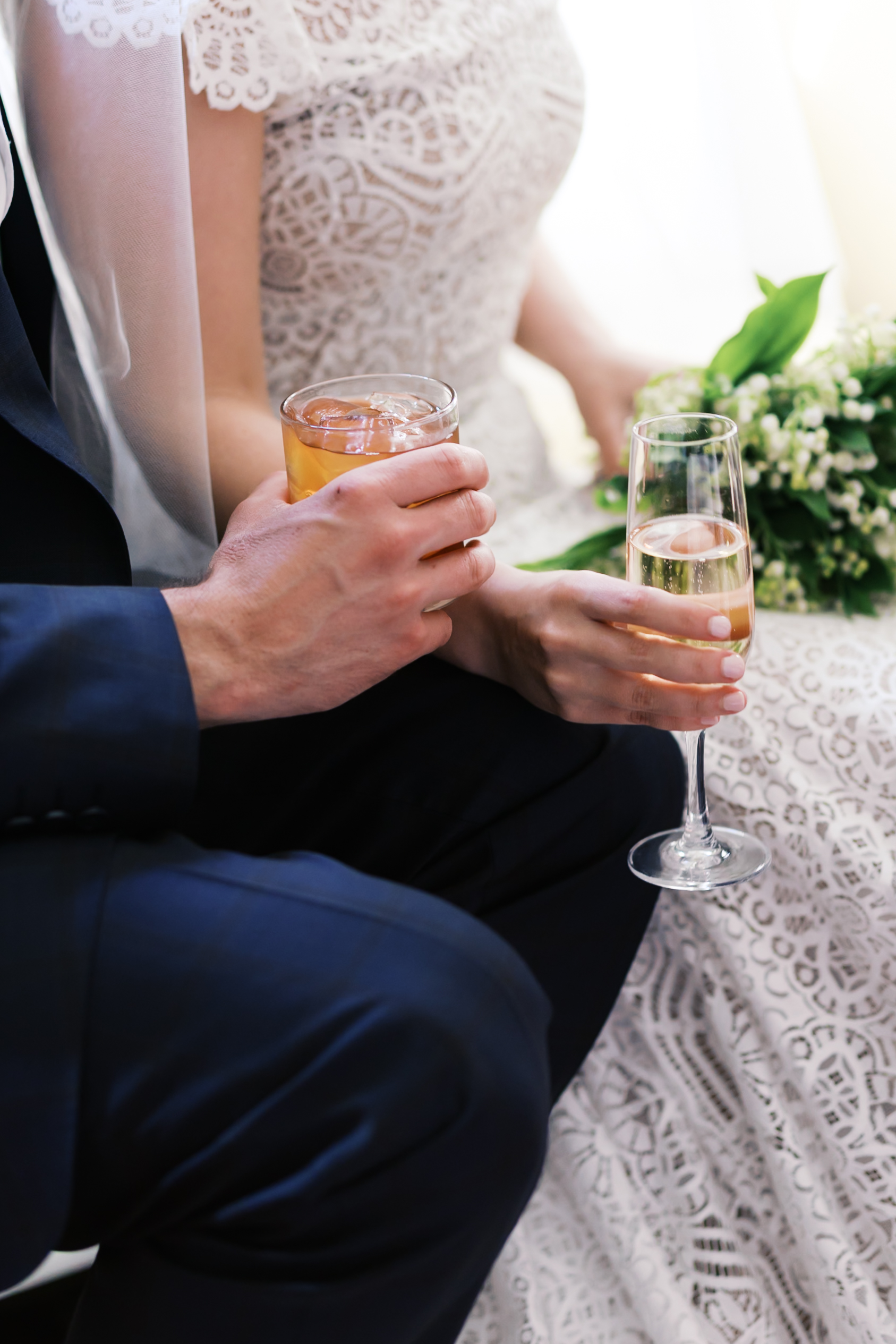 Close-up of bride and groom holding champagne and cocktail with bouquet in lap
