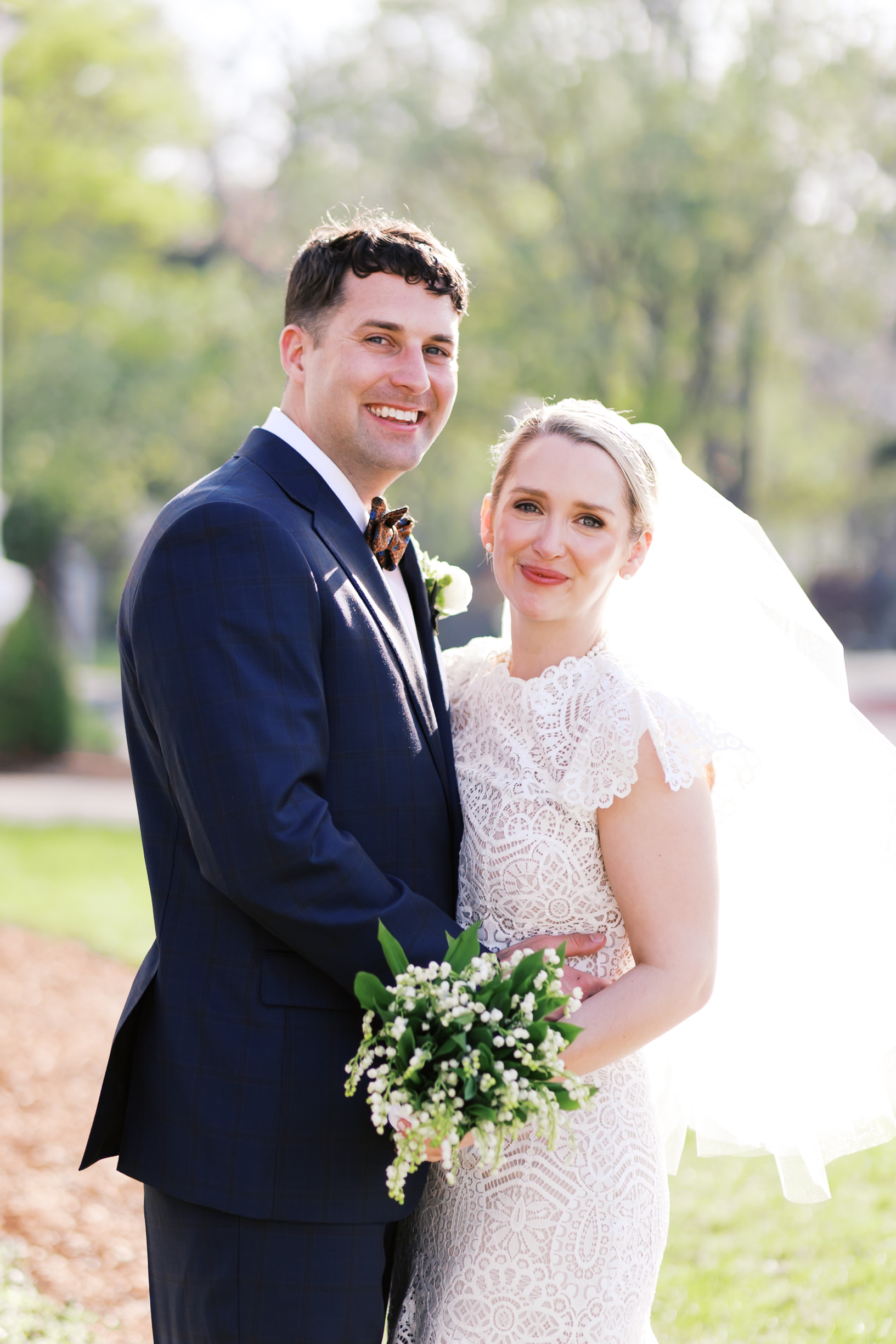 Bride and groom posing together outdoors, holding bouquet and smiling at camera