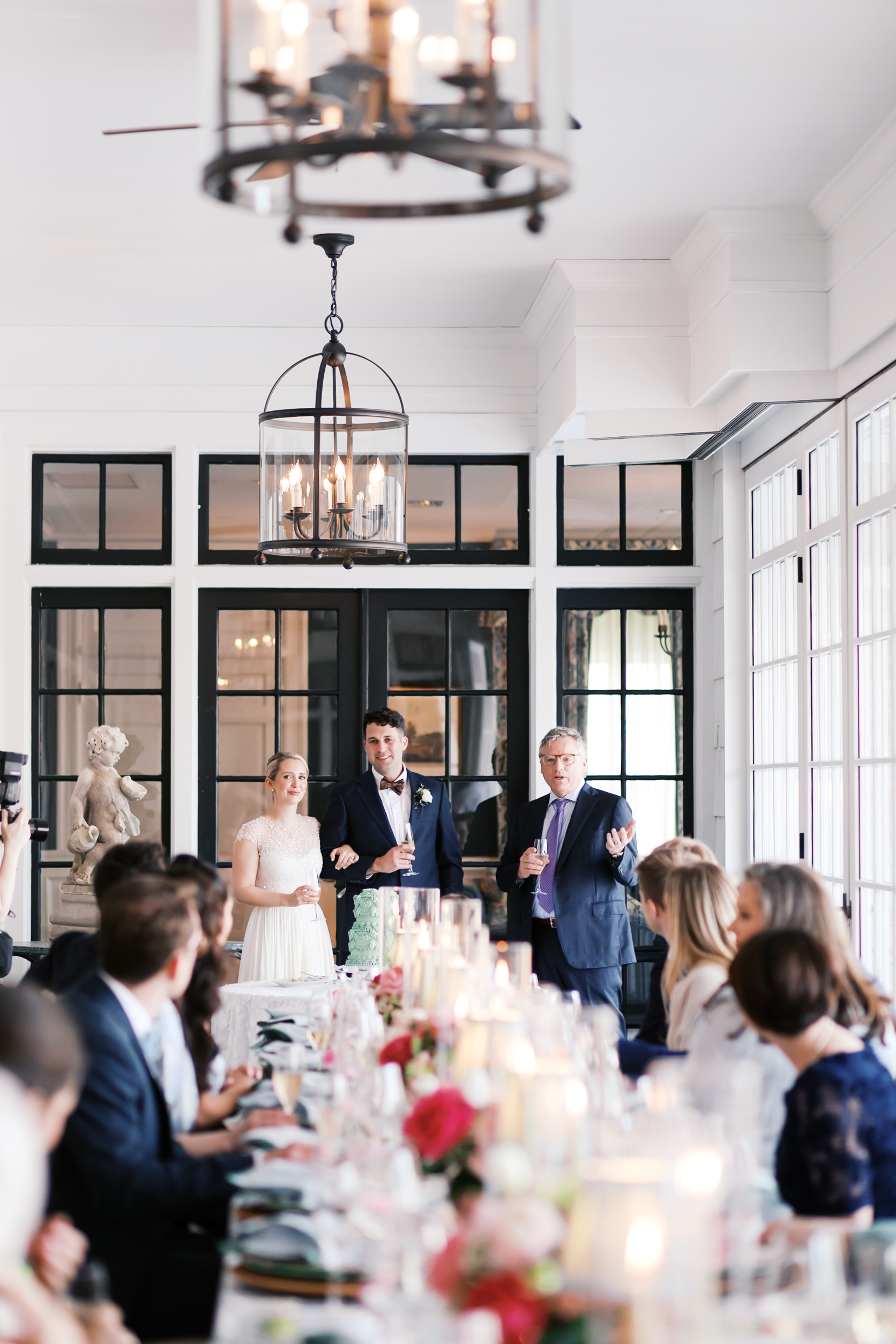 Bride and groom standing with guest giving speech during indoor wedding reception