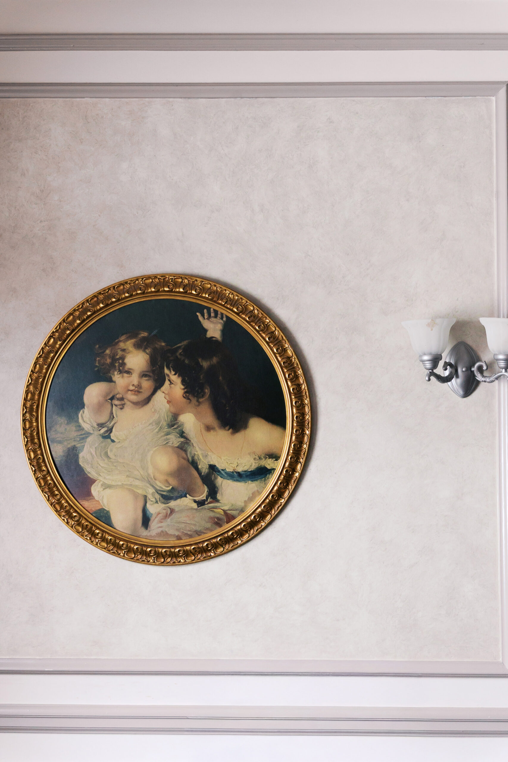 Round framed painting of two children hanging on a neutral wall beside a light fixture