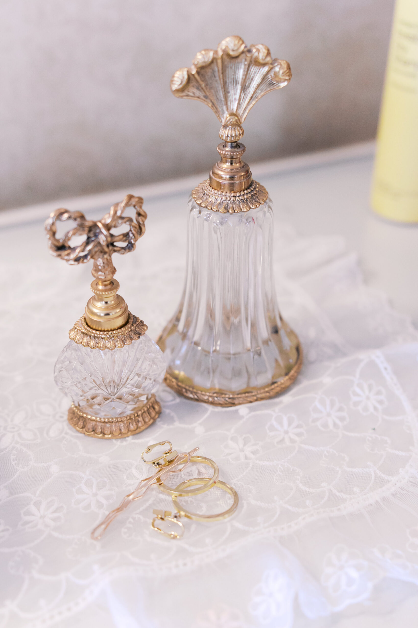 Two vintage-style glass perfume bottles with gold tops on lace fabric with jewelry nearby