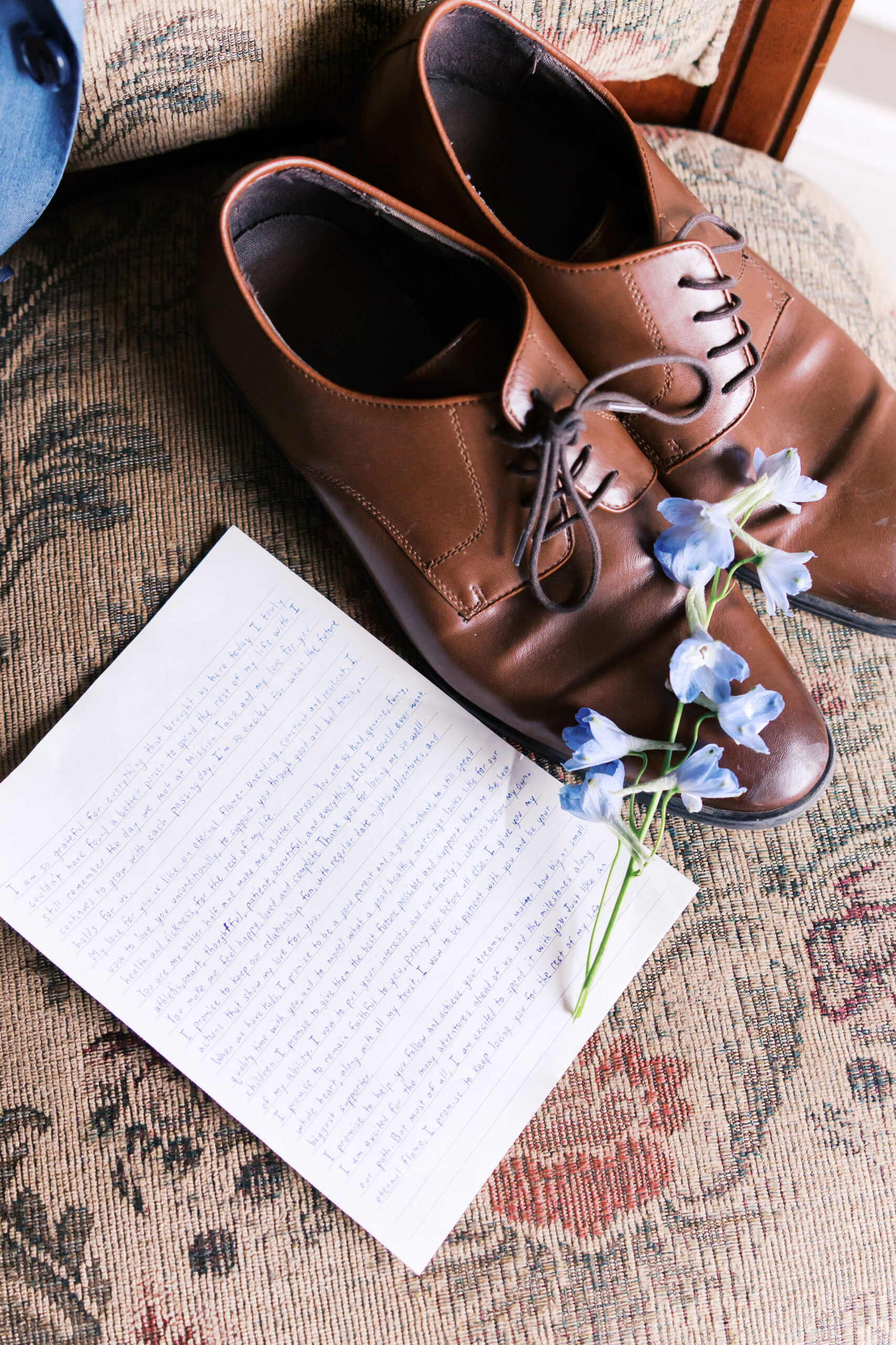 Brown leather dress shoes beside a handwritten letter and blue boutonniere on patterned chair