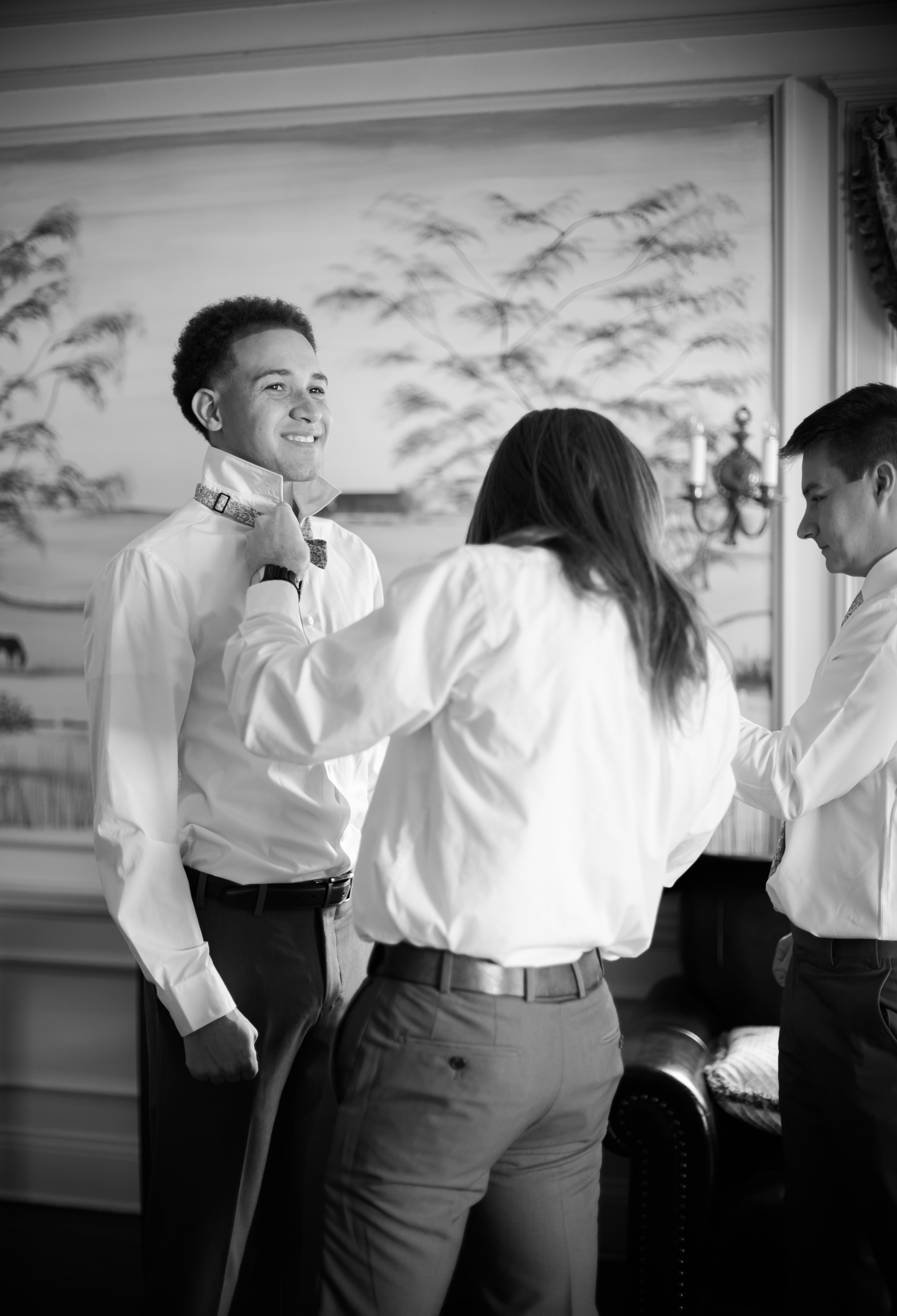 Two men helping groom adjust bow tie in softly lit room before ceremony