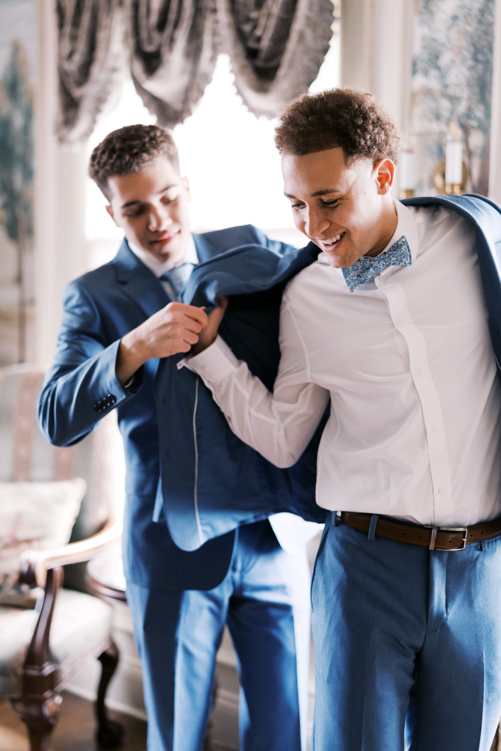 Man helping groom into blue suit jacket in bright room with window light
