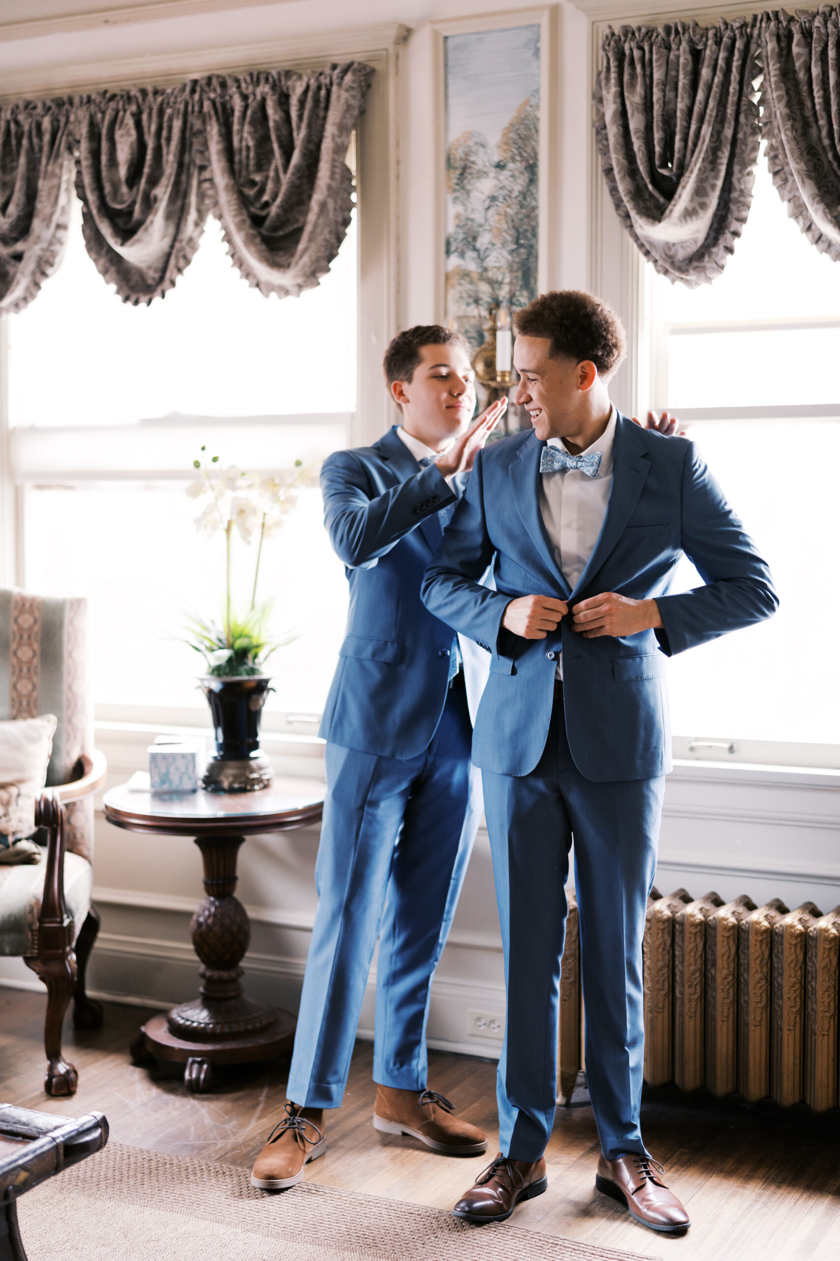 Man brushing off groom's blue suit jacket in bright room with window light