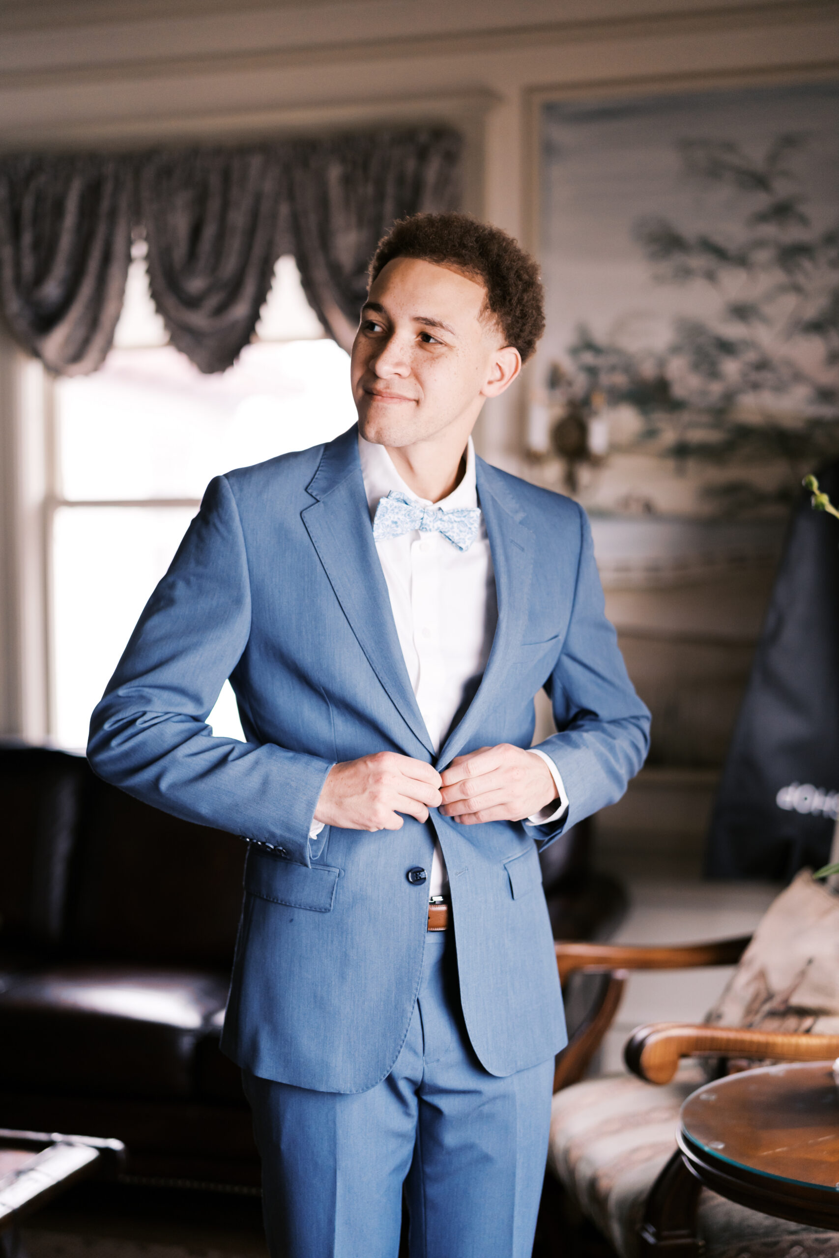 Groom buttoning blue suit jacket while standing in a warmly lit room