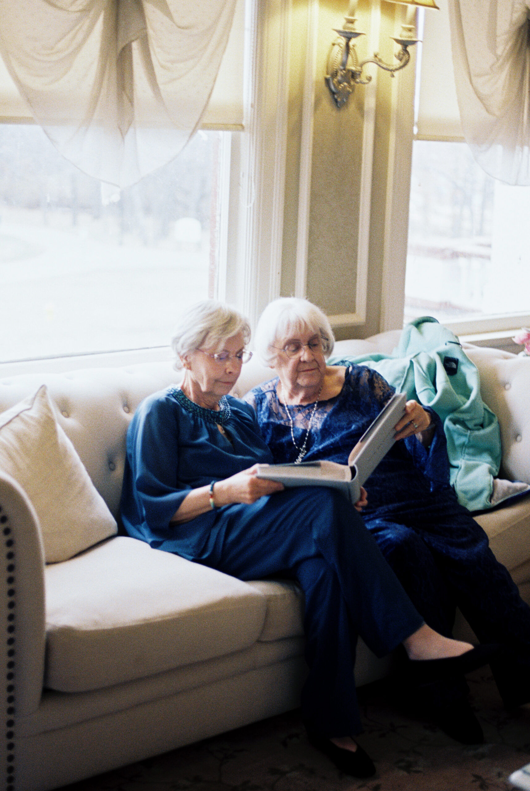 Two older women sitting on couch looking through a photo album by window light