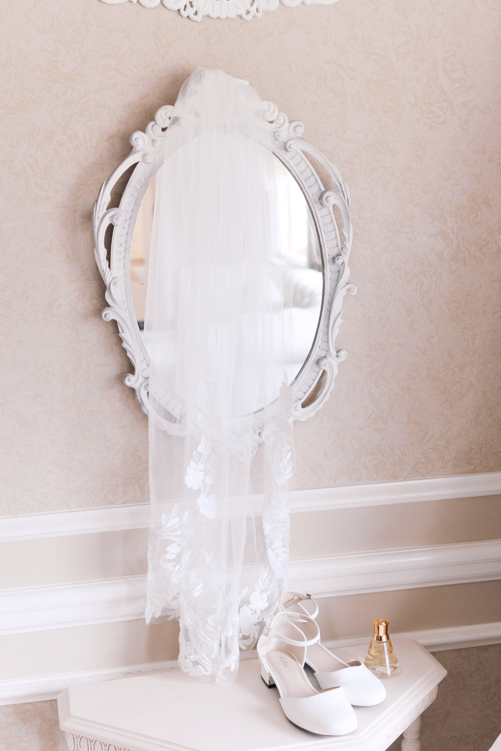 White bridal shoes, perfume bottle, and veil arranged beneath an ornate mirror