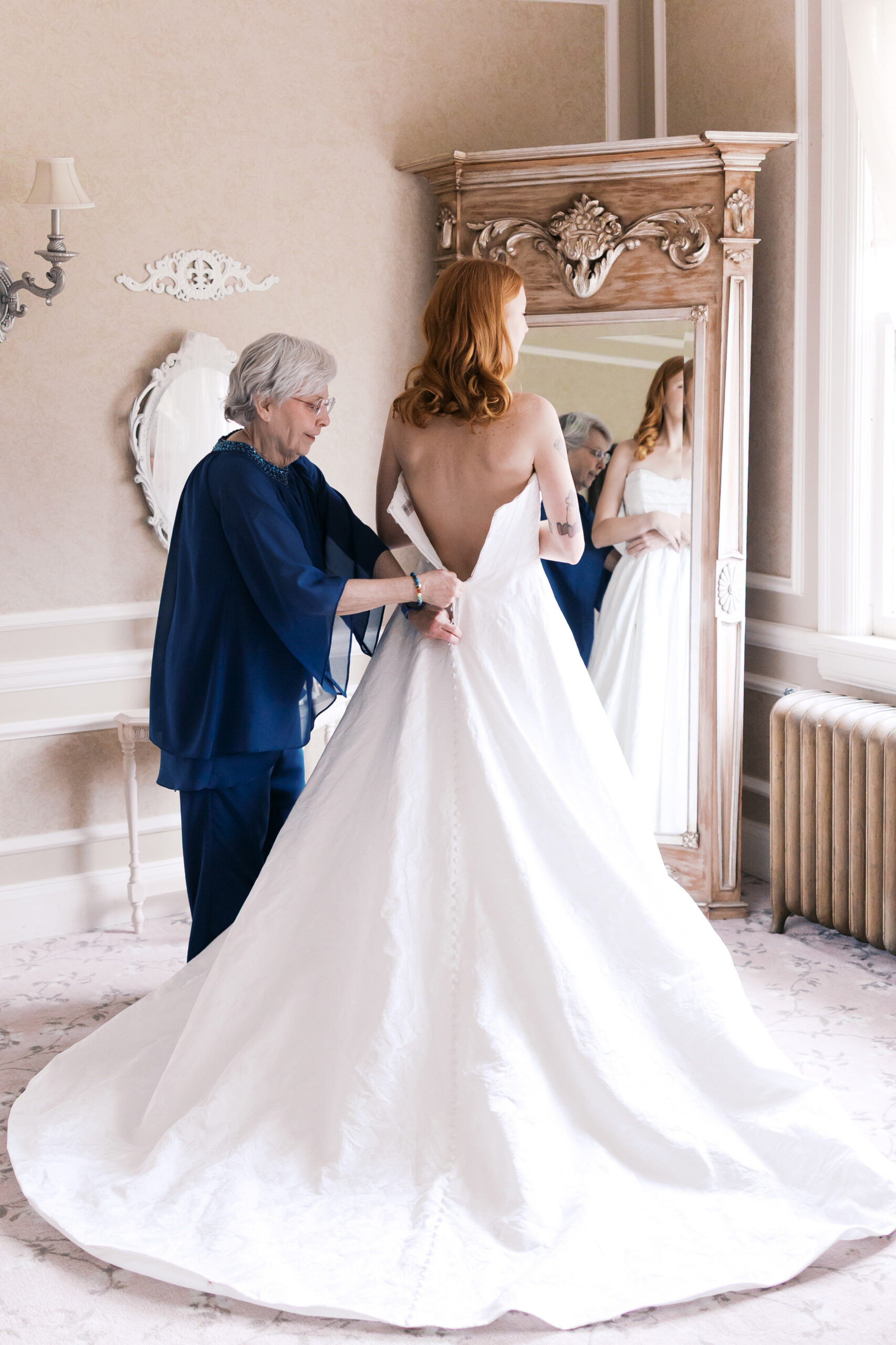 Bride in strapless wedding gown being buttoned by older woman in elegant Longview Mansion bridal suite