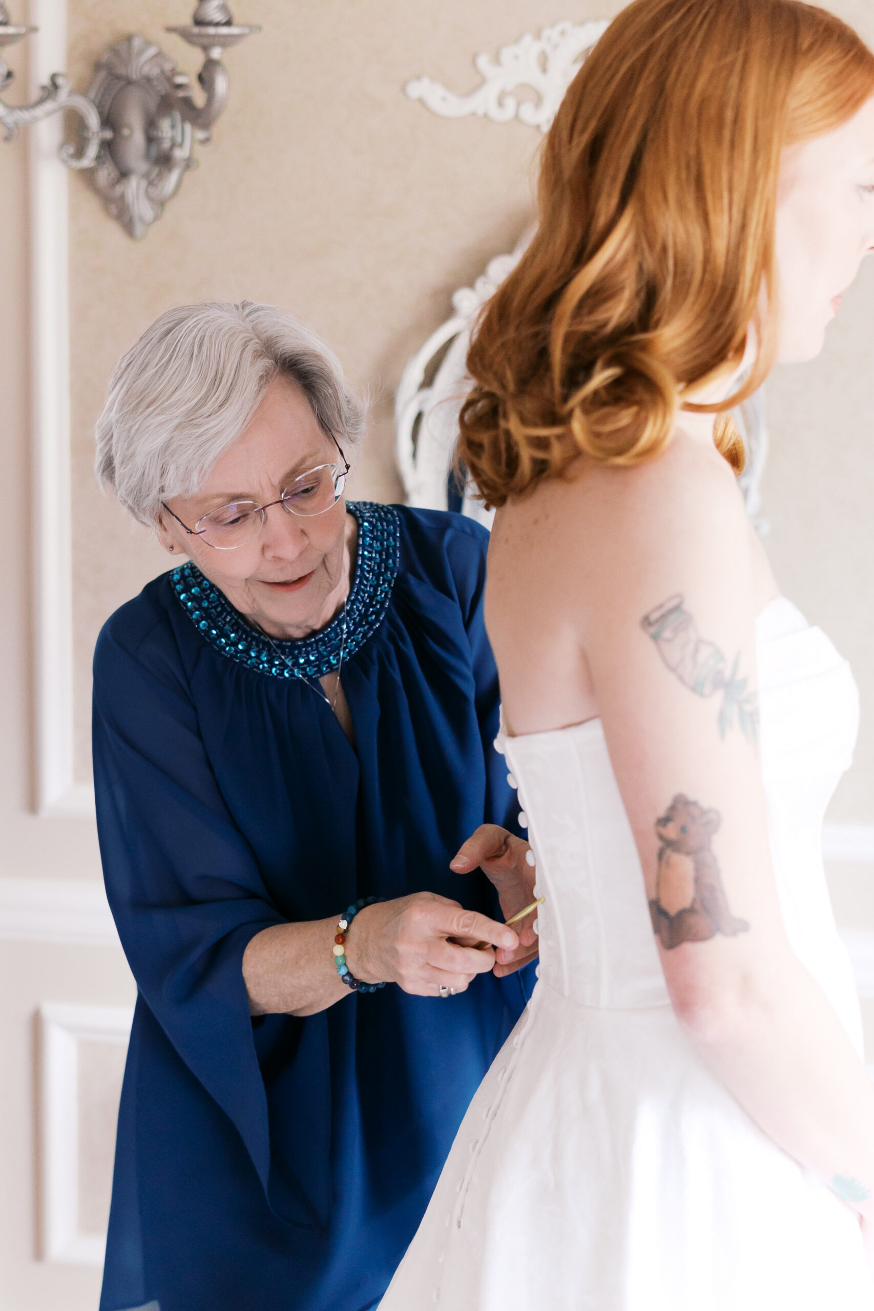 Close-up of older woman fastening bride’s wedding dress buttons in soft window light at Longview Mansion