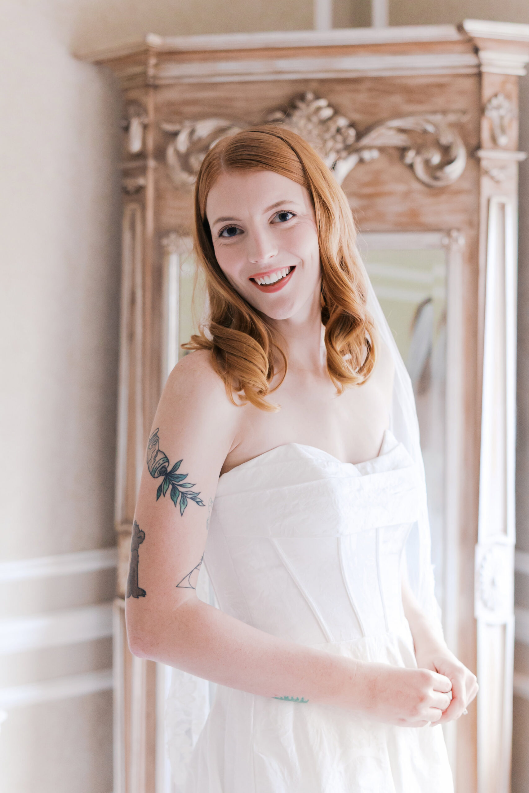 Bride smiling in strapless gown with veil, standing in front of ornate mirror