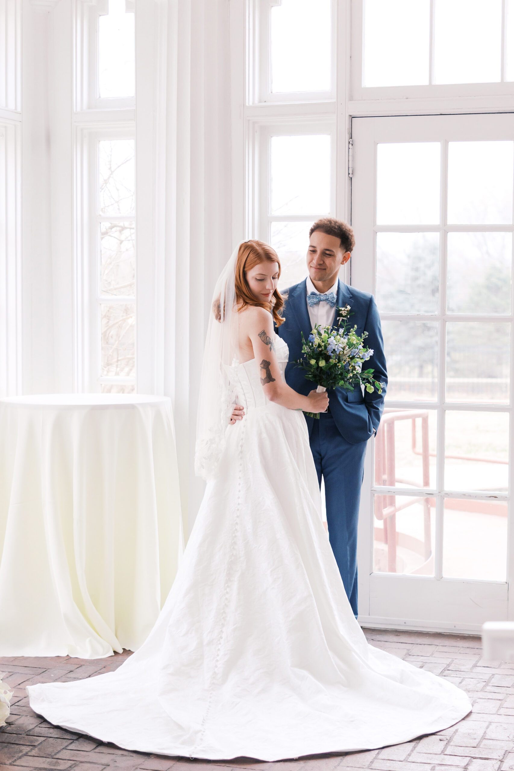 Bride and groom posing by tall windows with soft light and bouquet