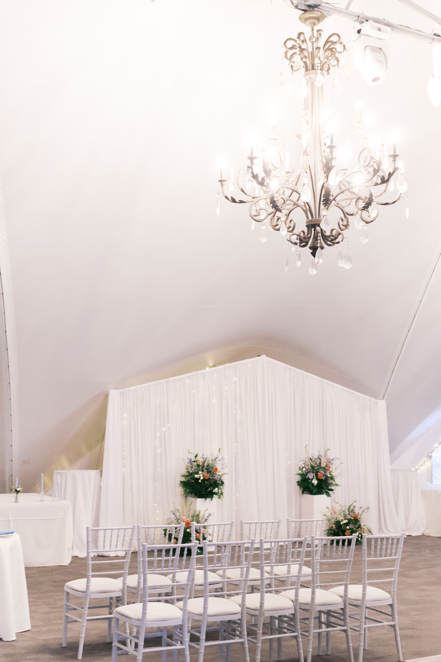 Elegant indoor ceremony setup with white chairs, floral arrangements, and chandelier