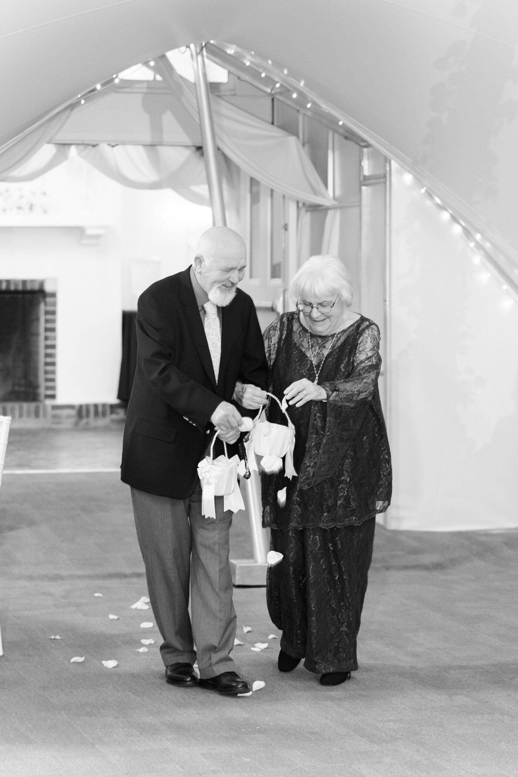 Elderly couple in formal attire scatter flower petals down aisle inside softly lit wedding tent.