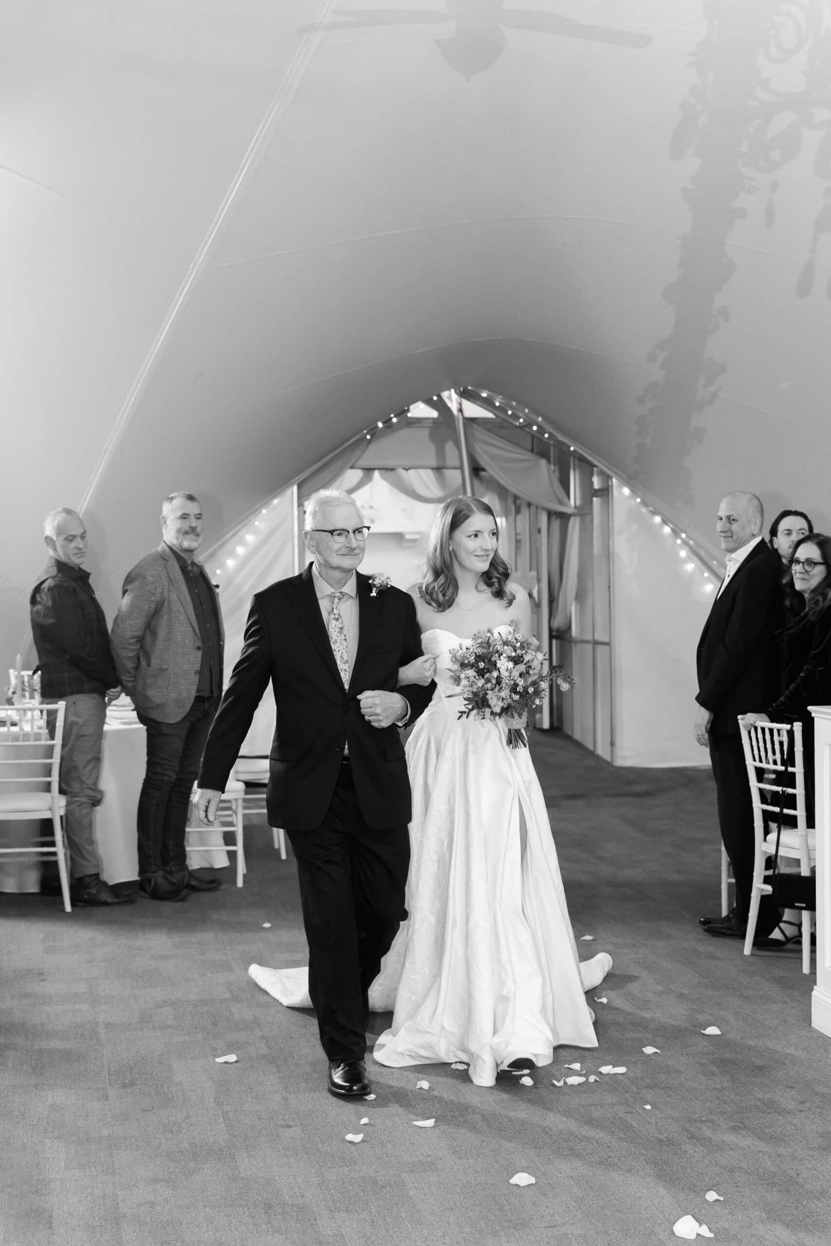 Bride walks down aisle with older man, guests standing on either side inside elegant tent venue.