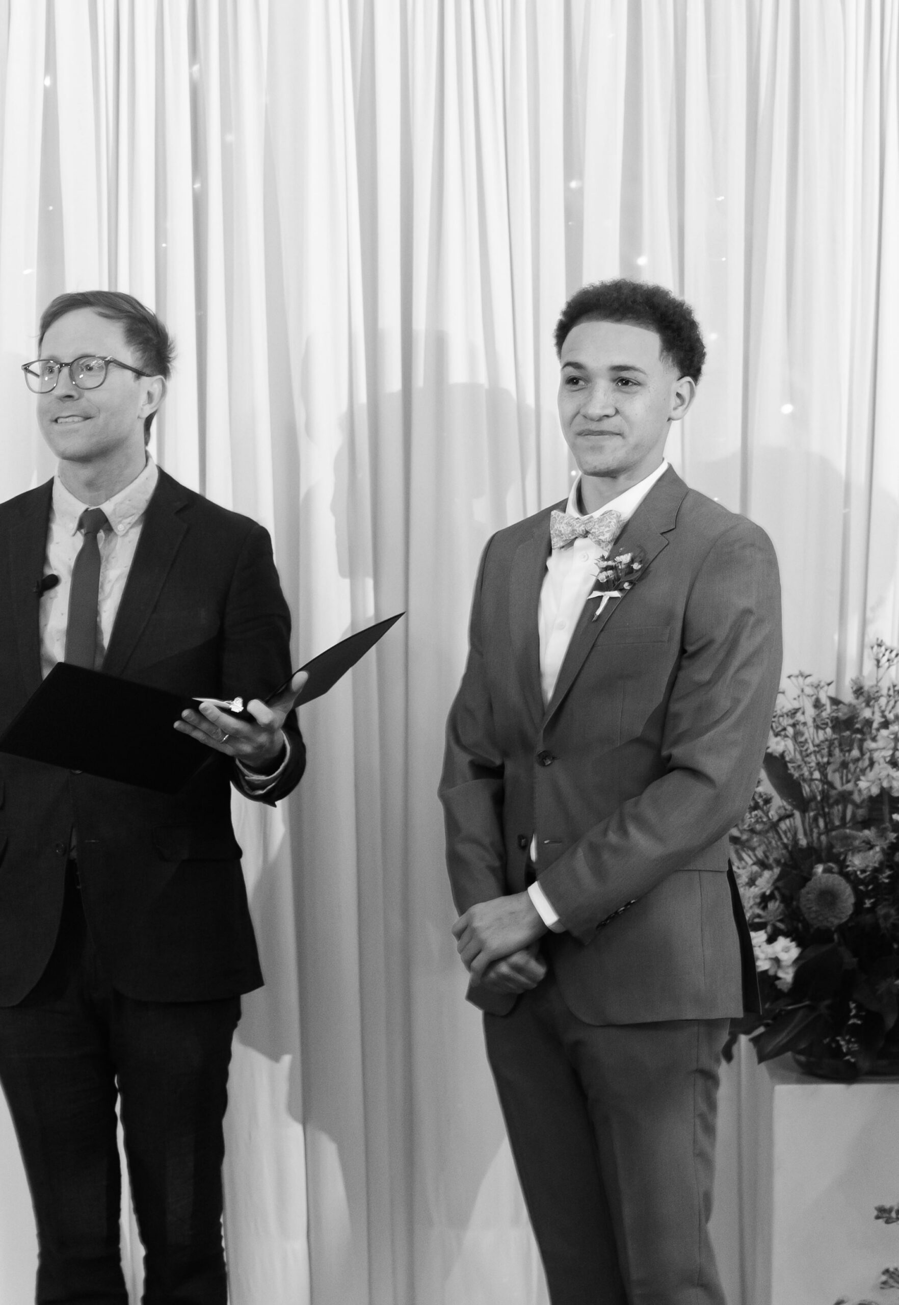 Groom stands beside officiant holding folder during indoor wedding ceremony with draped backdrop.