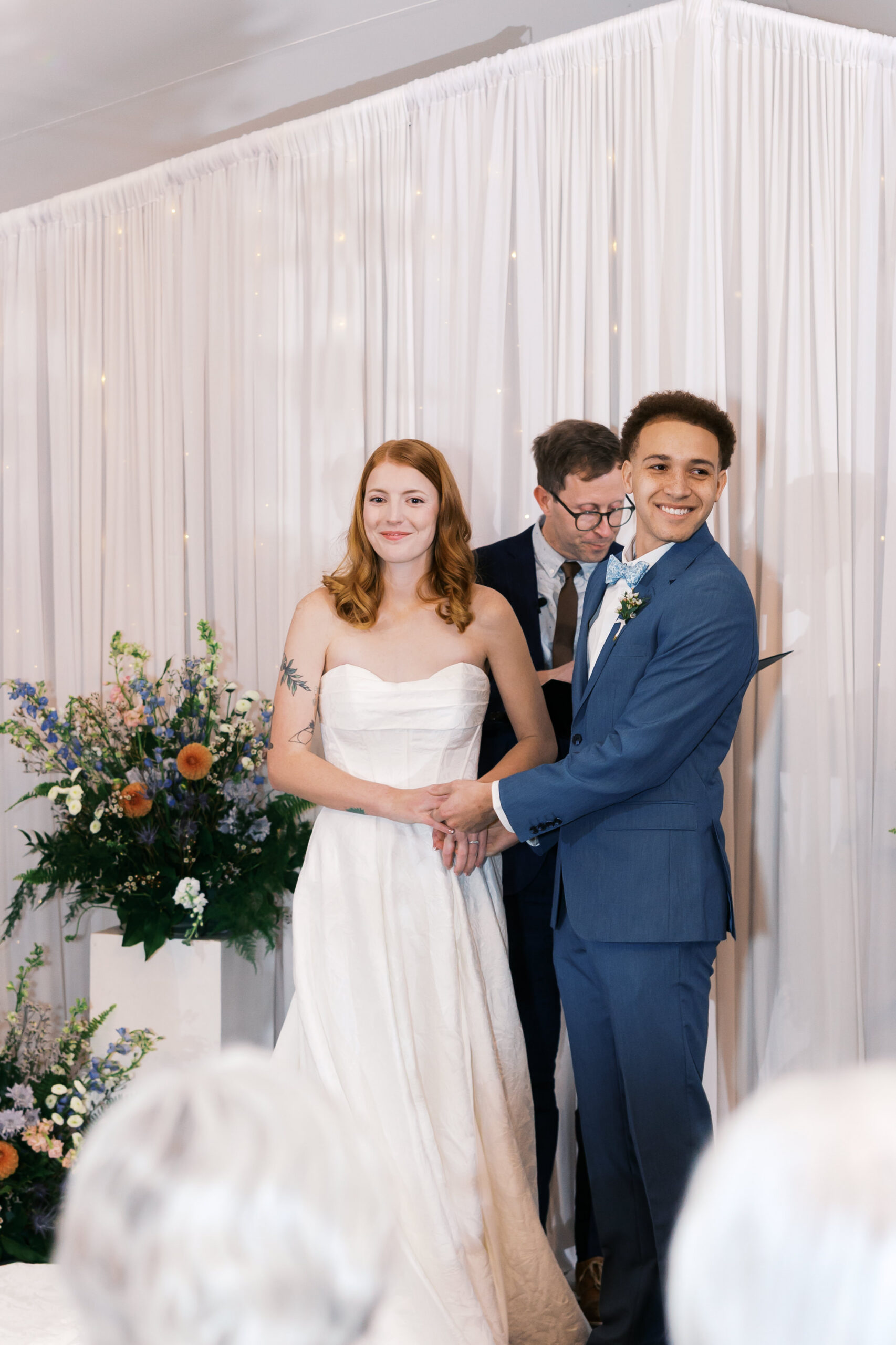 Bride and groom hold hands at altar during ceremony, officiant reading behind them.