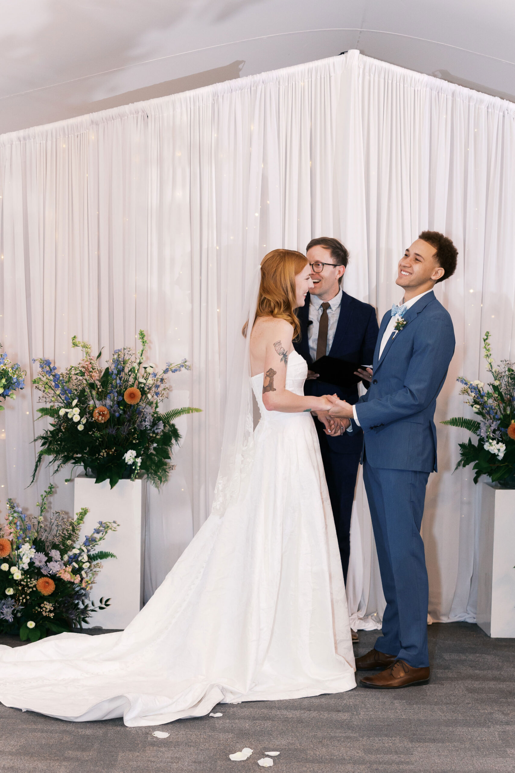 Bride and groom face each other holding hands during ceremony with white draped backdrop and florals.