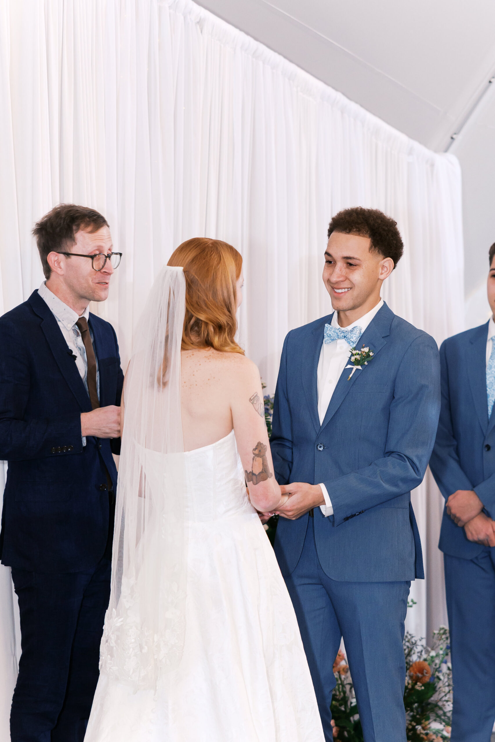 Groom smiles at bride as they exchange vows, officiant speaking beside them.
