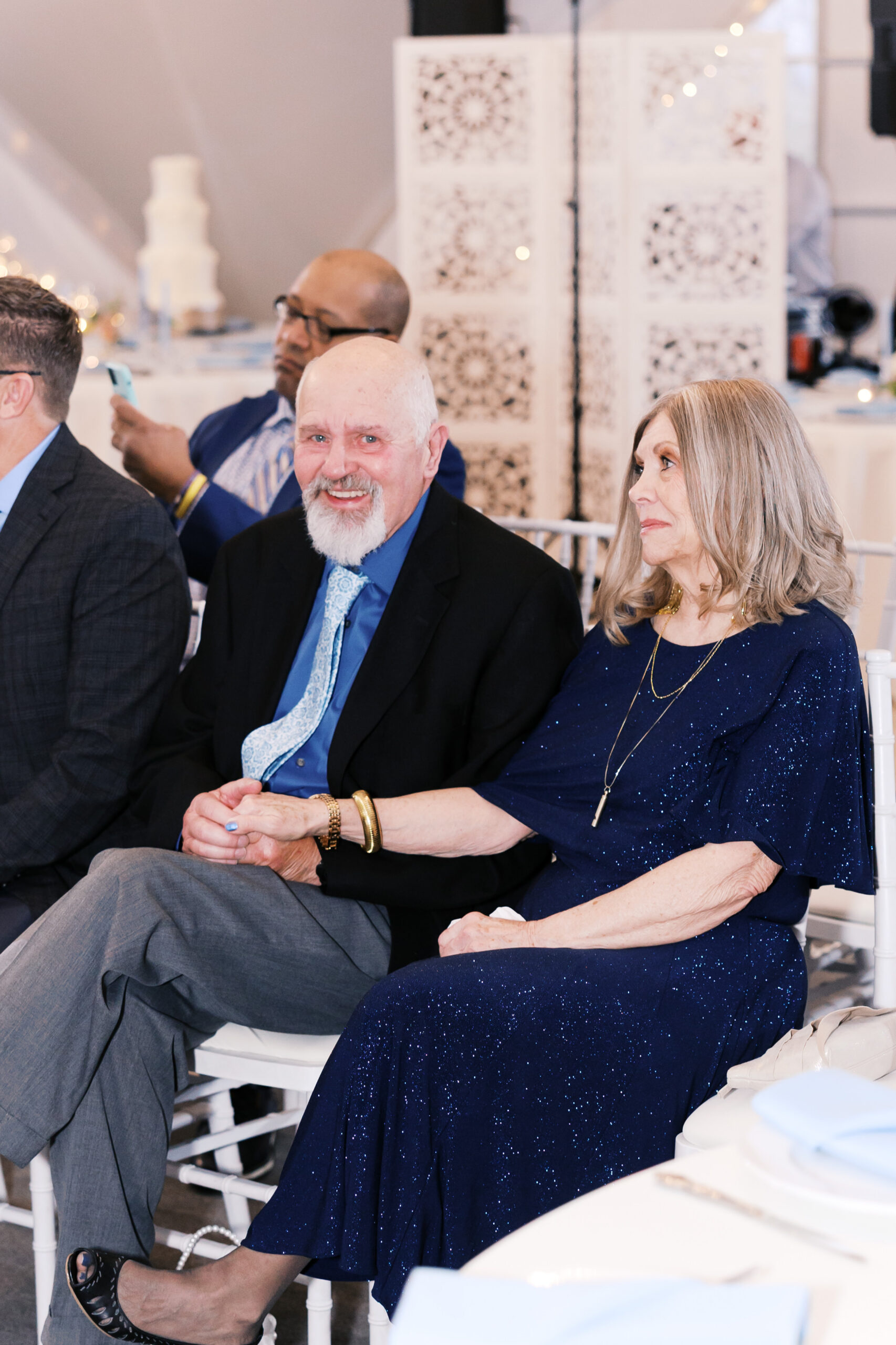 Older couple seated together at ceremony, holding hands and watching with soft smiles.