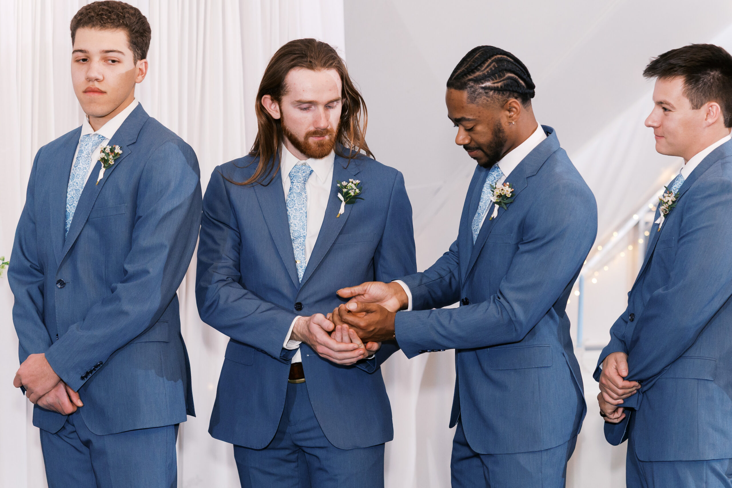 Groomsmen in matching blue suits stand together as one hands another the rings.