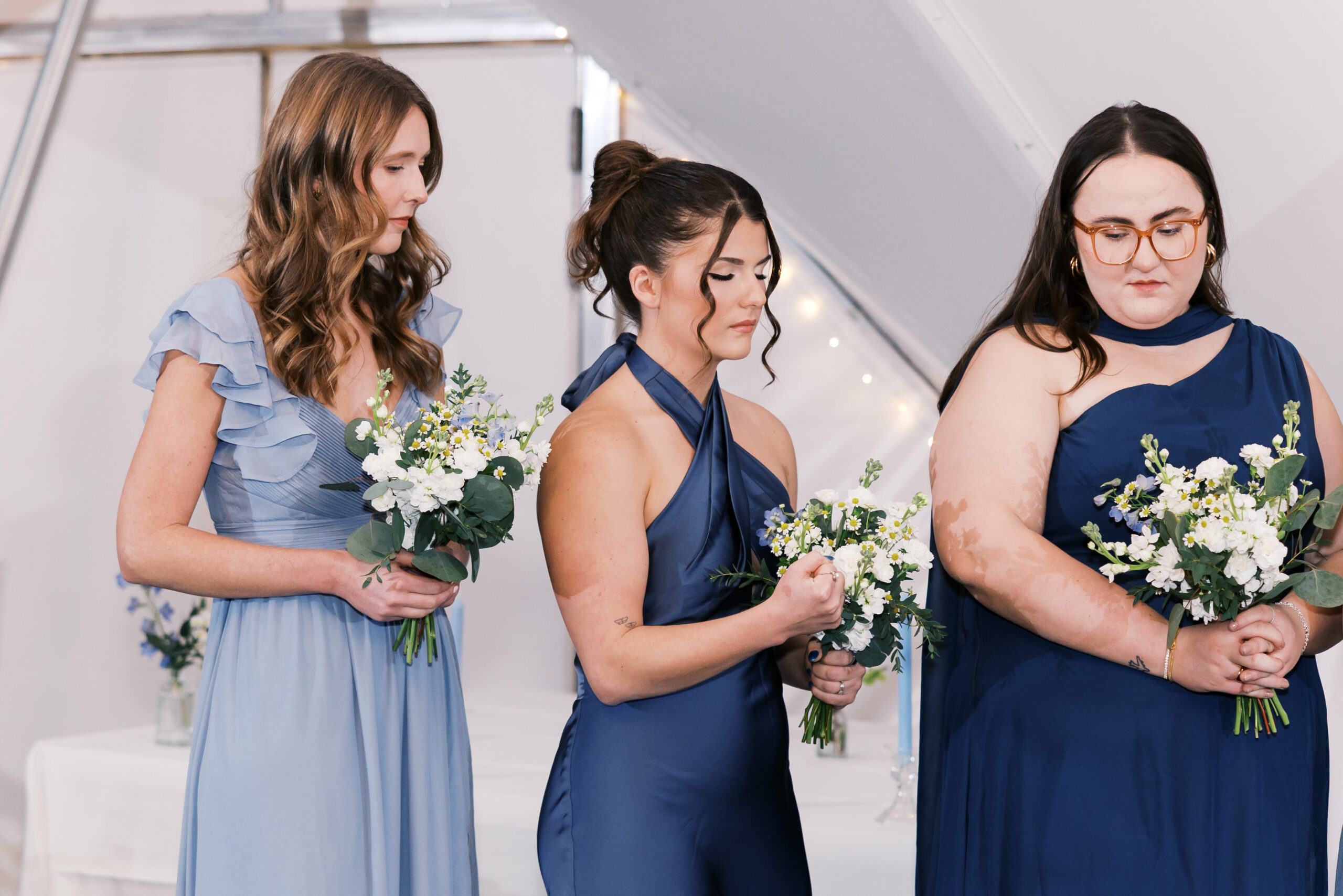 Bridesmaids in blue dresses hold white and green bouquets while standing during indoor wedding ceremony.