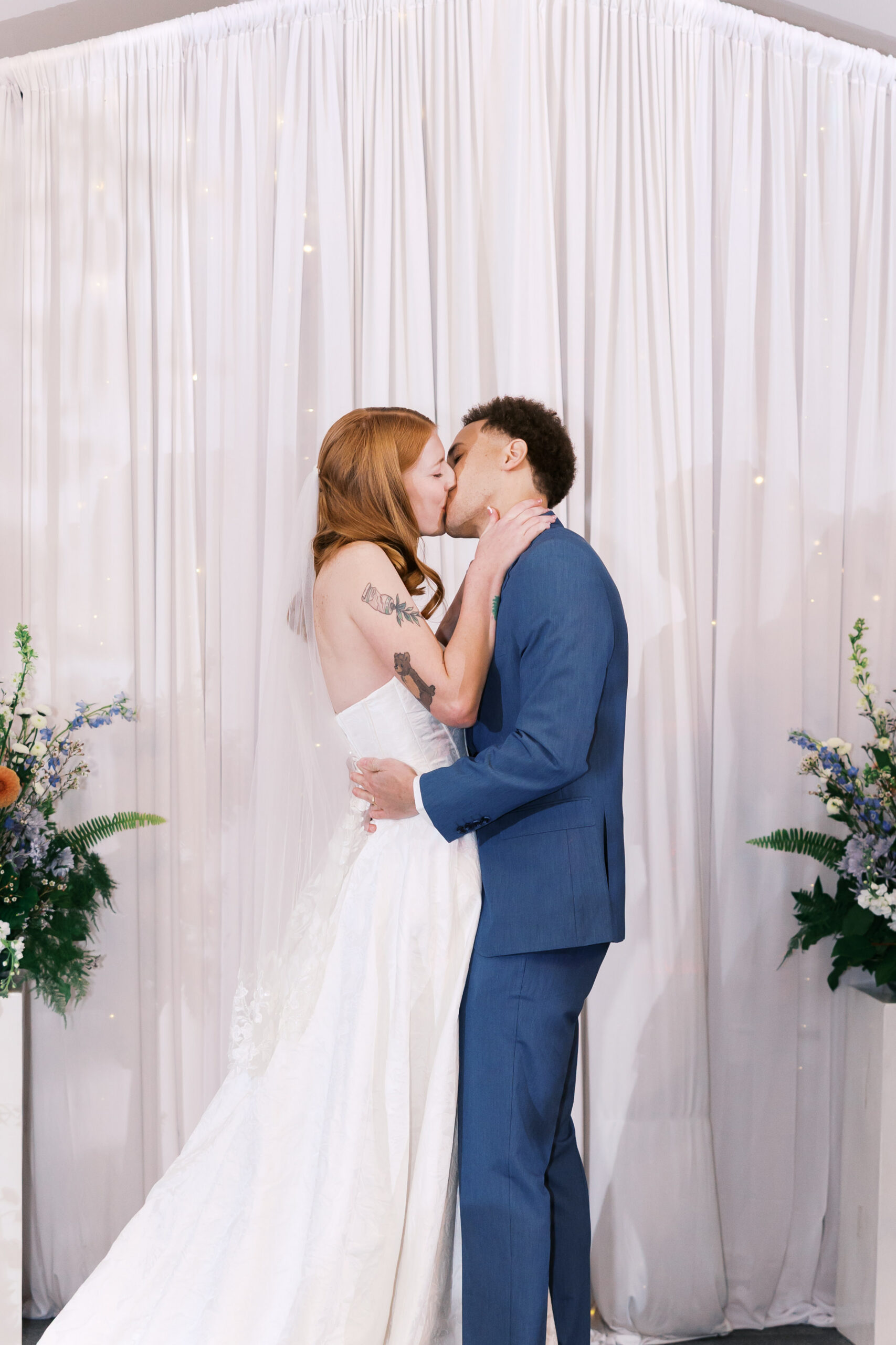 Bride and groom share a kiss at the altar with floral arrangements and white drapery backdrop.