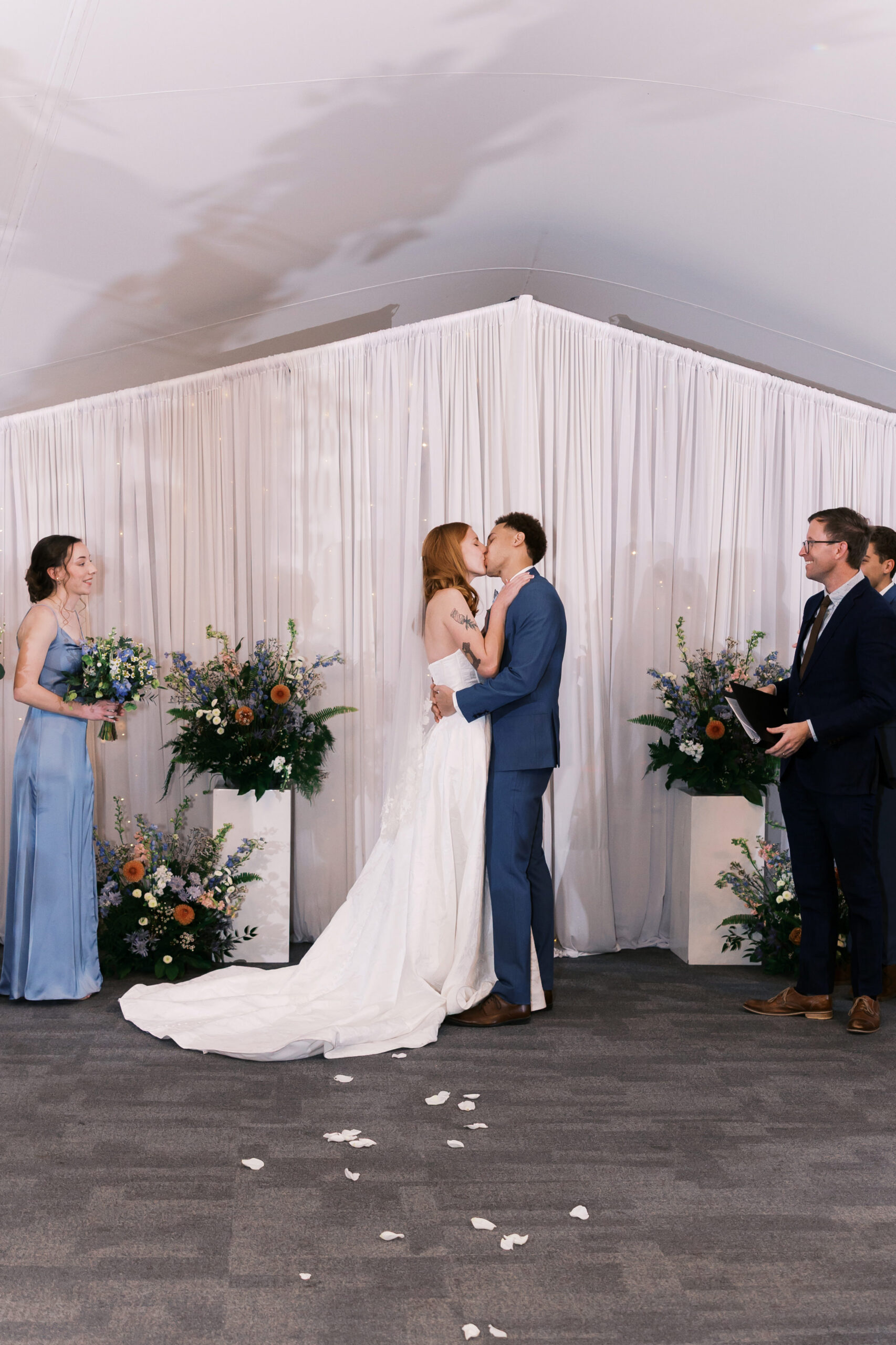 Wide view of indoor wedding ceremony as couple kisses at altar surrounded by florals and guests.