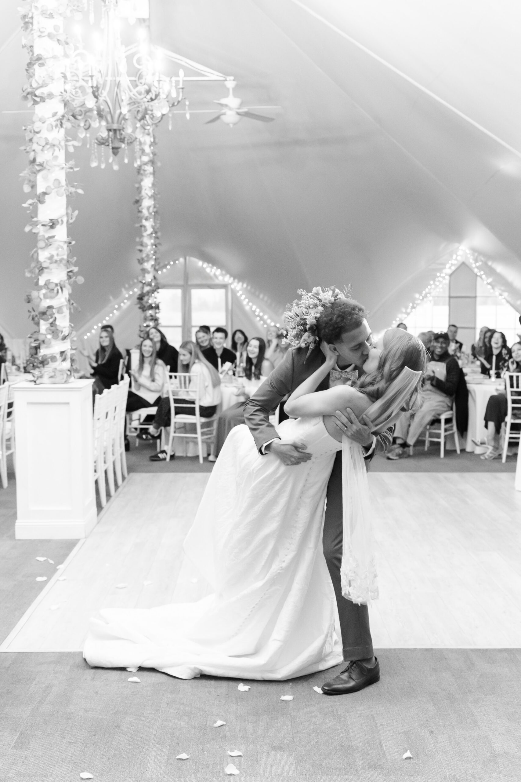Bride and groom kiss during reception dance as guests watch under chandeliers in elegant venue.