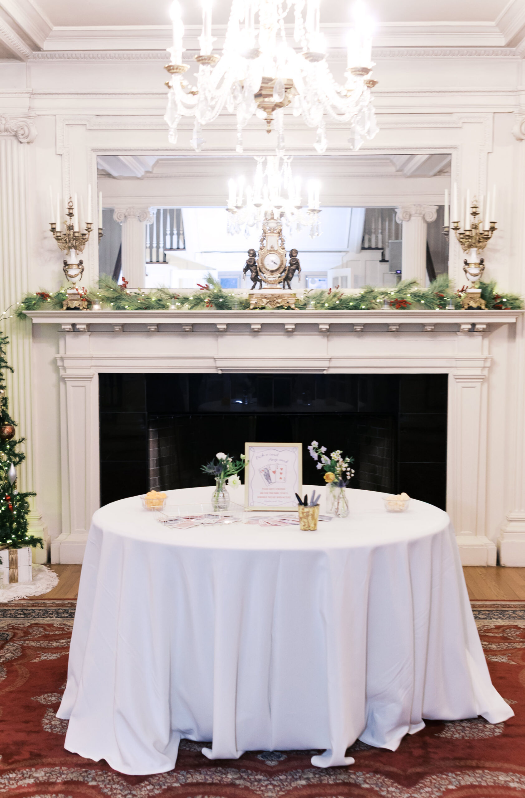 Wedding reception detail with round table, fireplace mantel, greenery garland, and chandelier above.