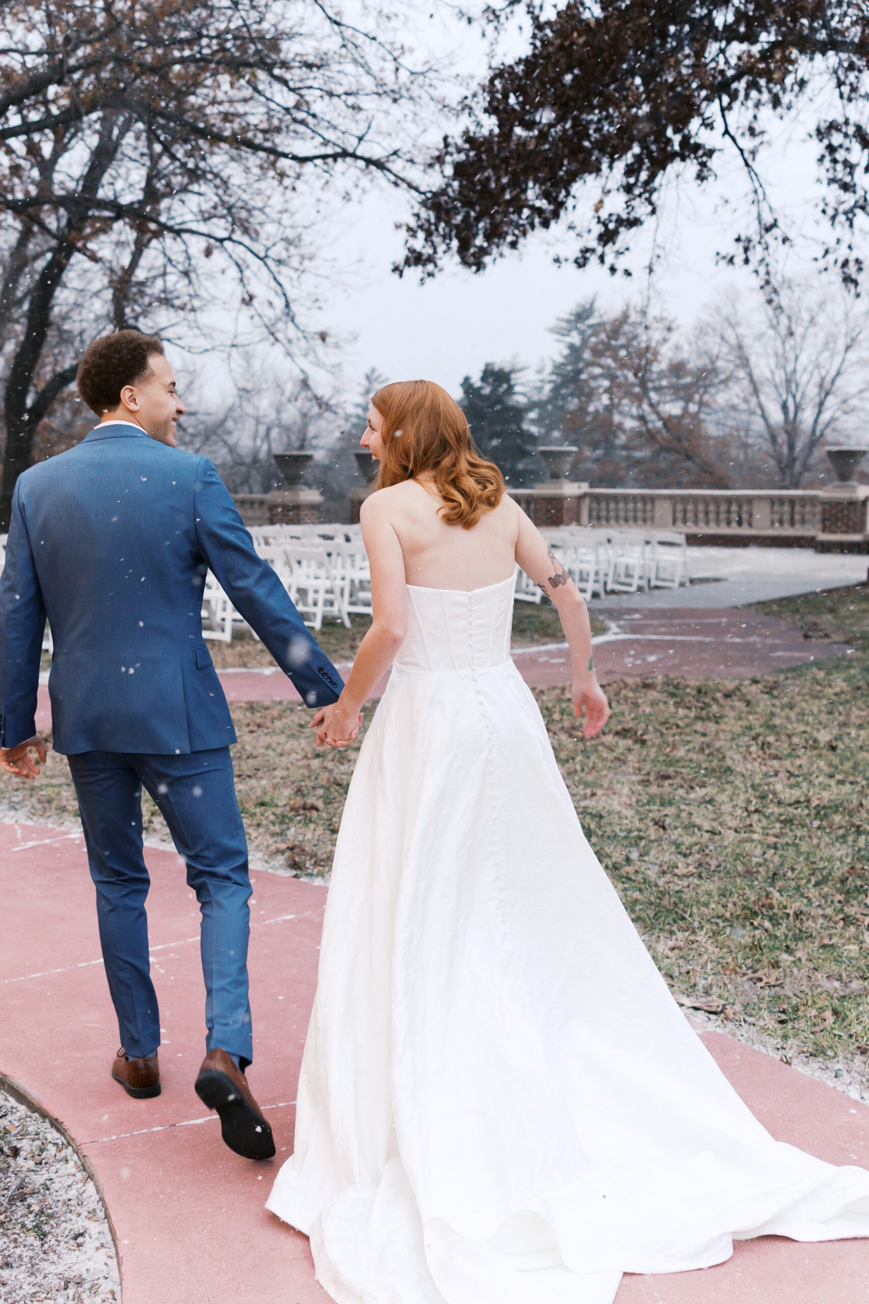 Bride and groom walk hand in hand outdoors in light snowfall after ceremony at winter wedding venue.