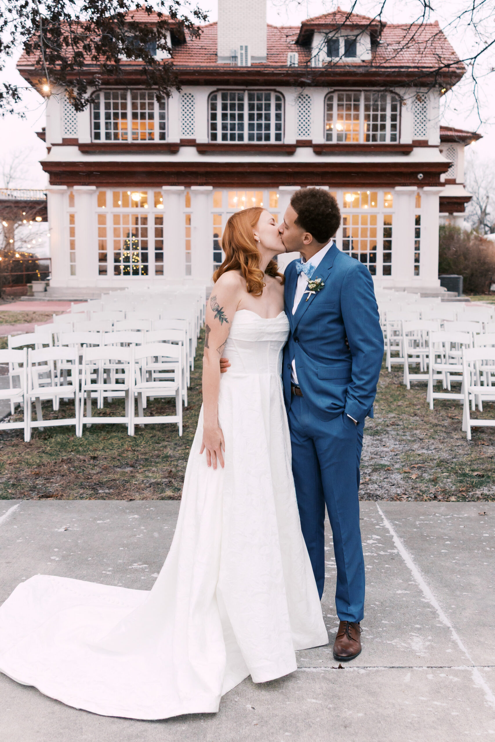 Bride and groom kiss in front of large historic building with rows of white ceremony chairs outside.