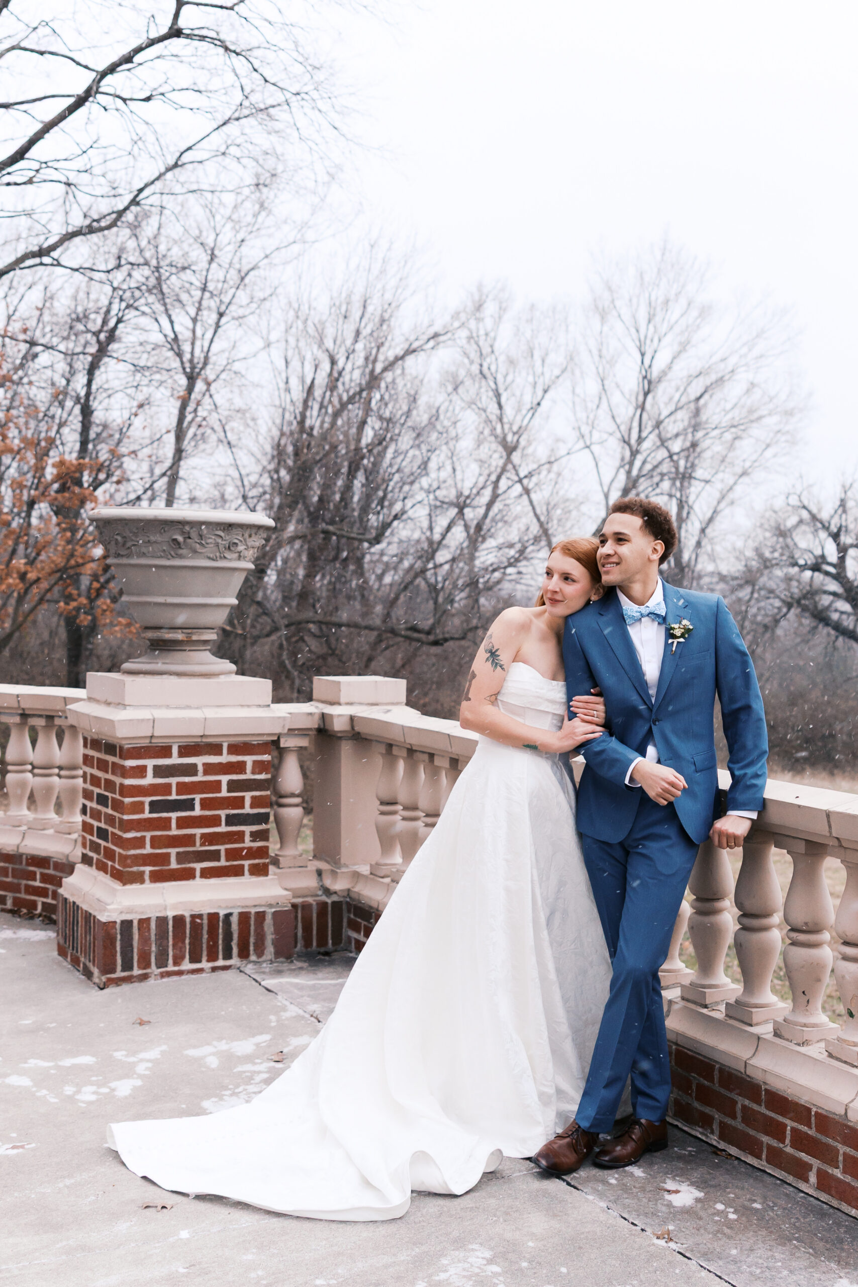 Newlyweds stand together on stone terrace, embracing during light snowfall with trees in background.