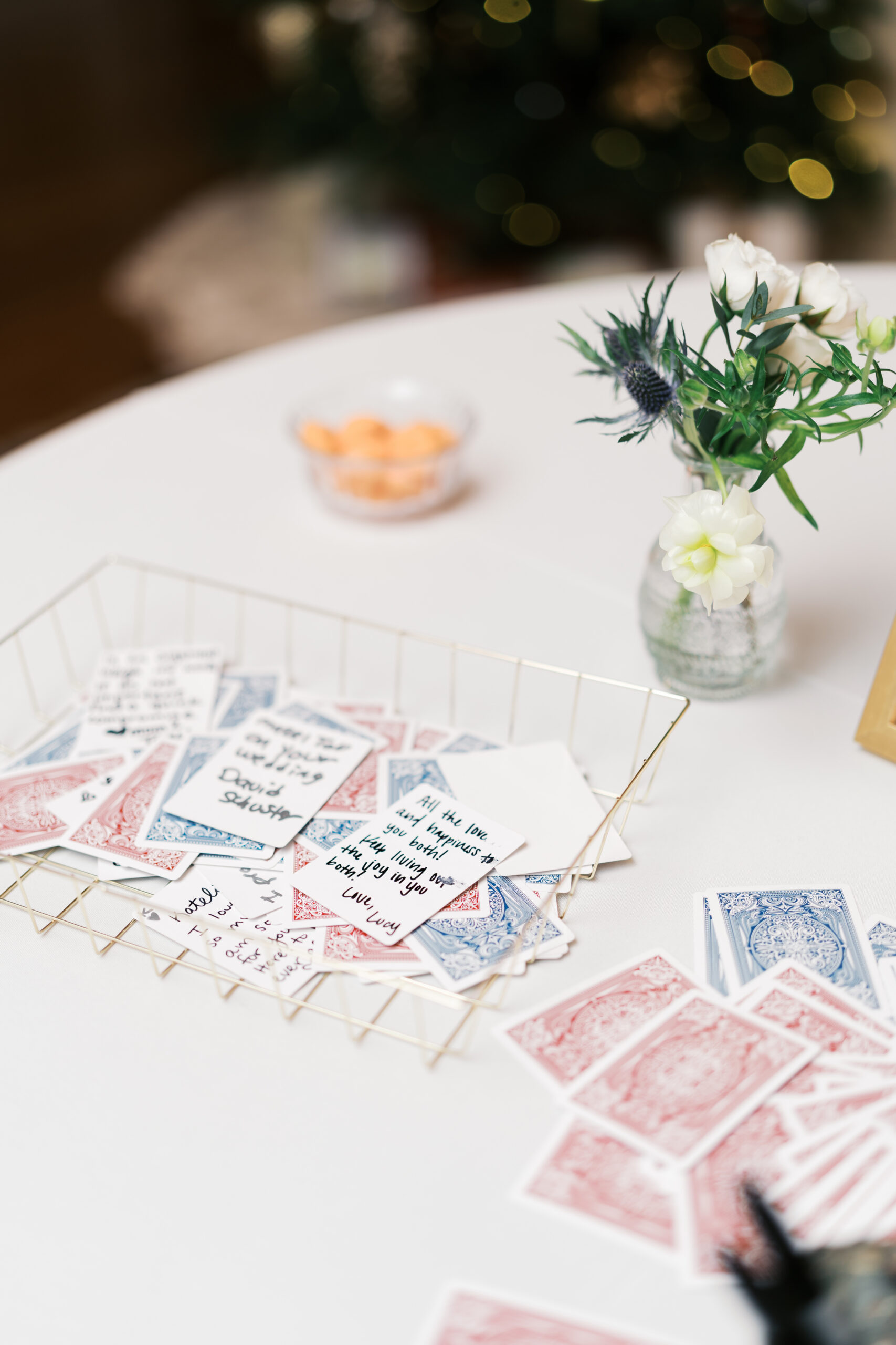 Playing cards with messages written on them on a table with a small flower arrangement and white tablecloth.
