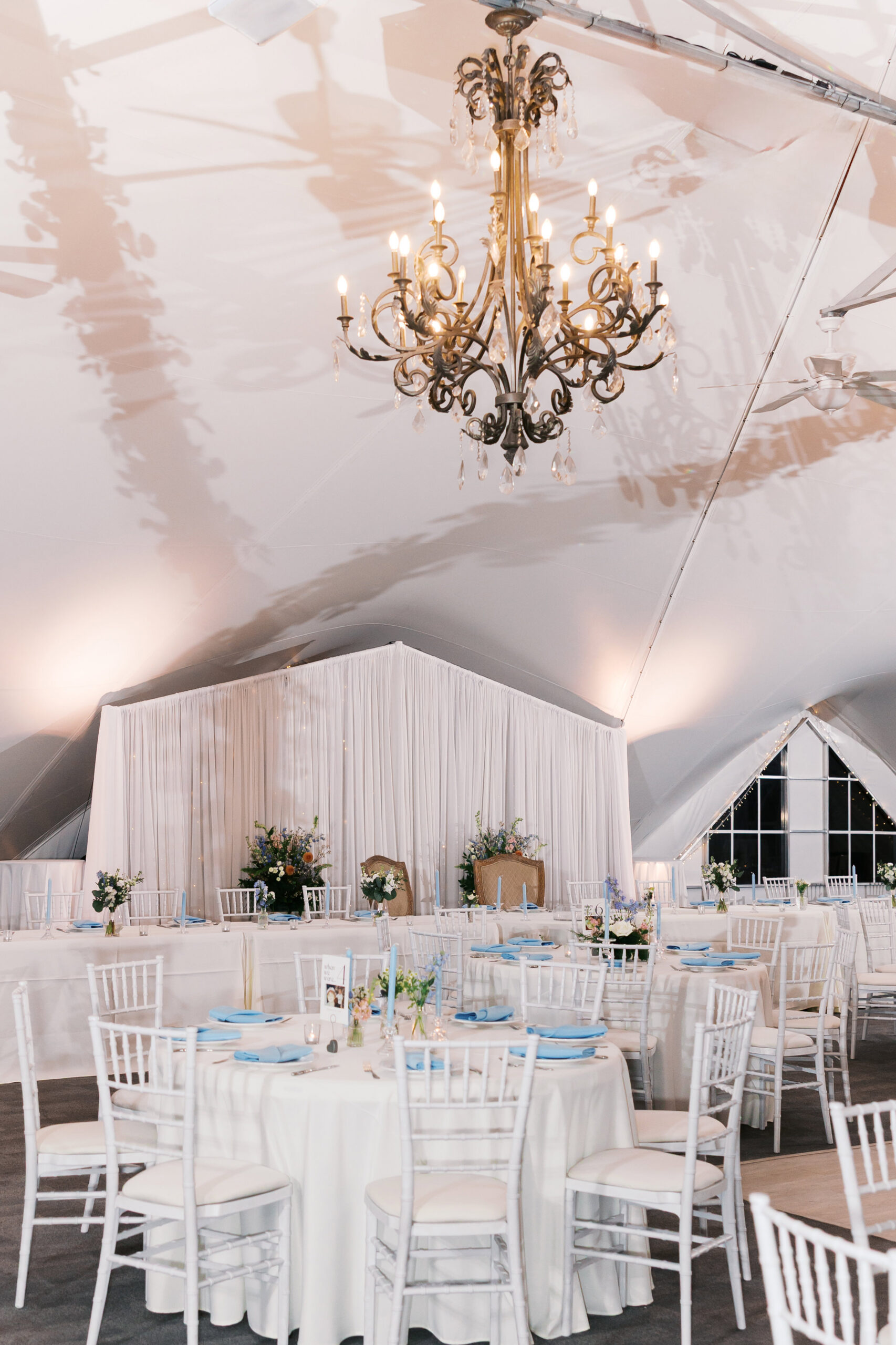 Elegant wedding reception tent with chandeliers, round tables, white linens, and blue napkin accents