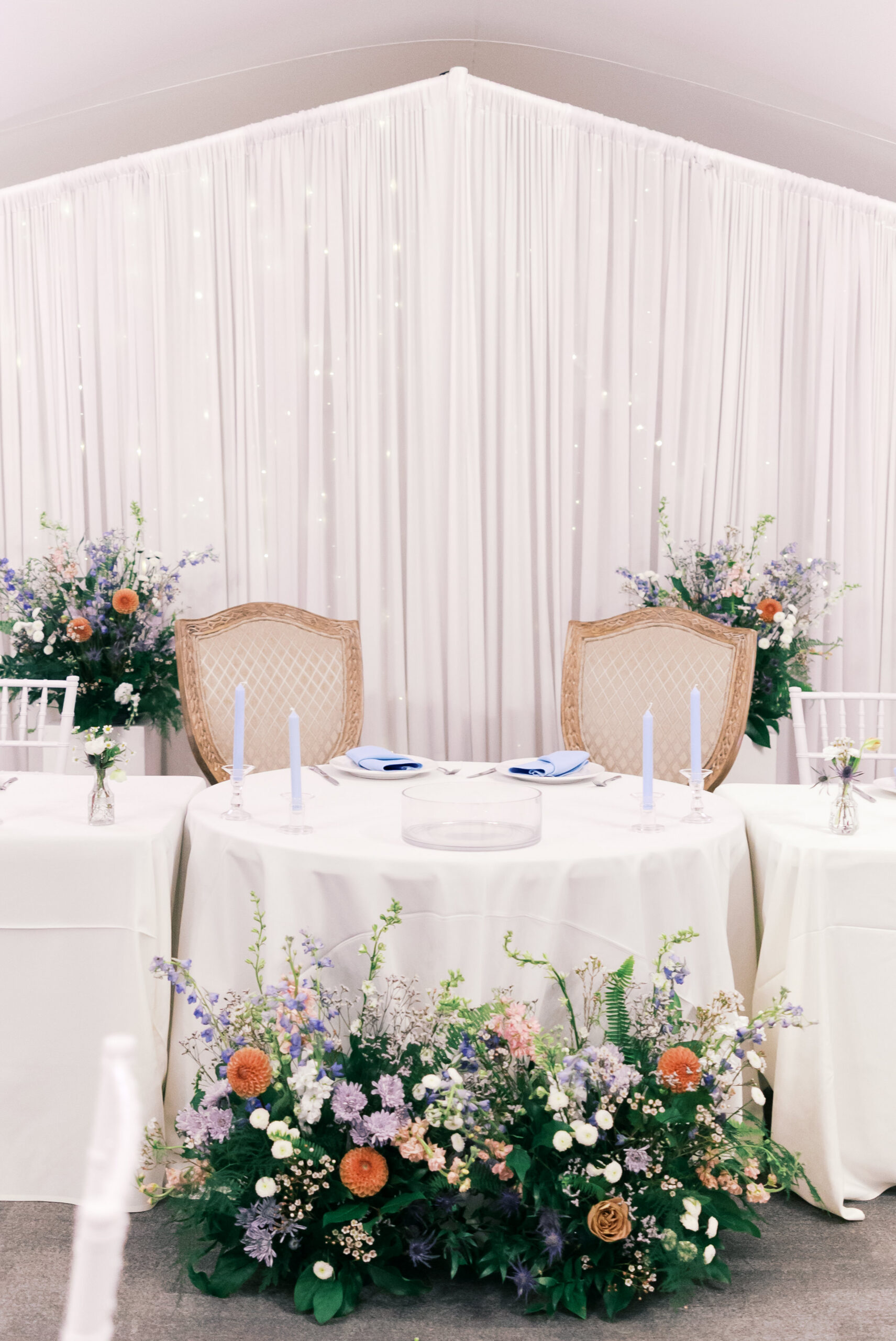 Sweetheart table with white draping backdrop, pastel floral arrangements, and blue taper candles