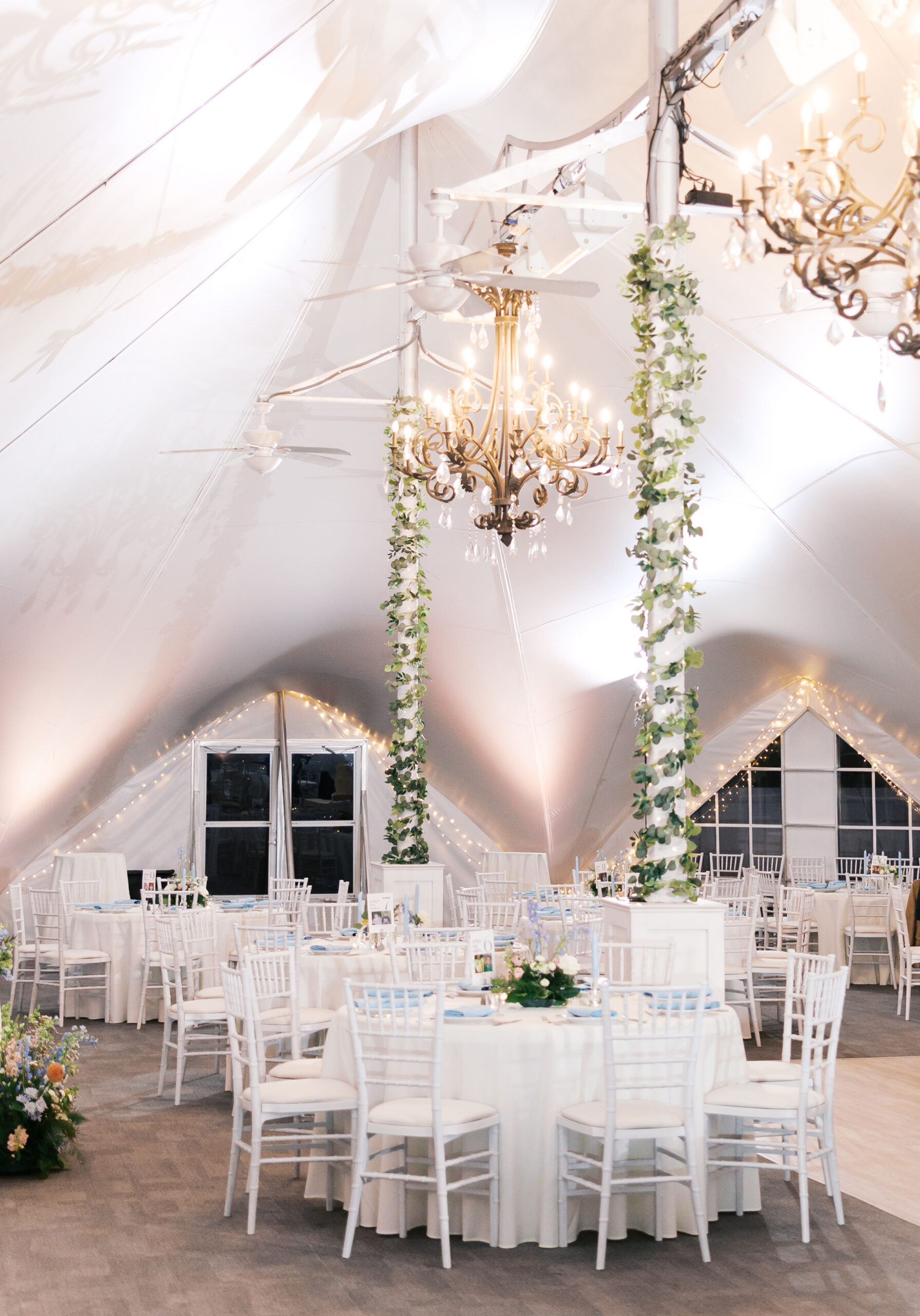 Wedding tent interior with chandeliers, greenery-wrapped columns, and round tables set for reception