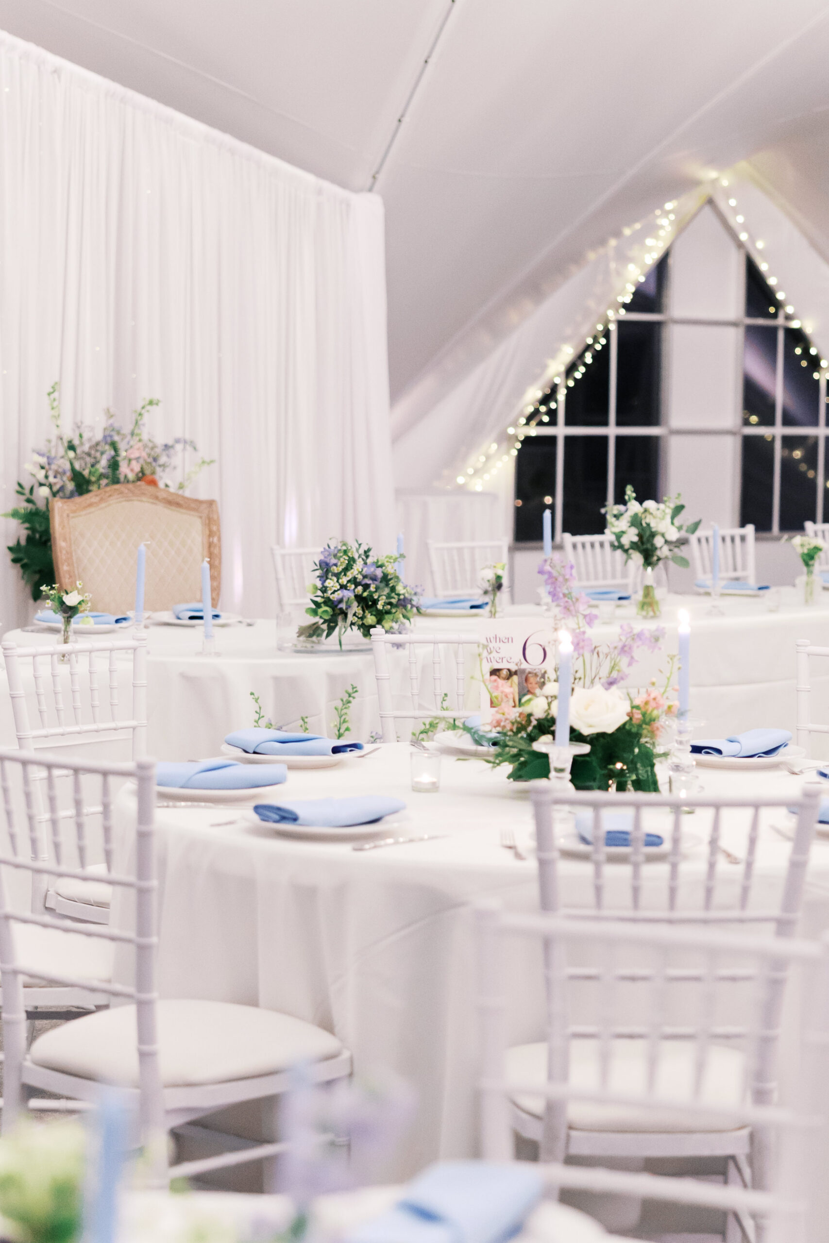Reception tables with white linens, blue napkins, floral centerpieces, and string lights in tent