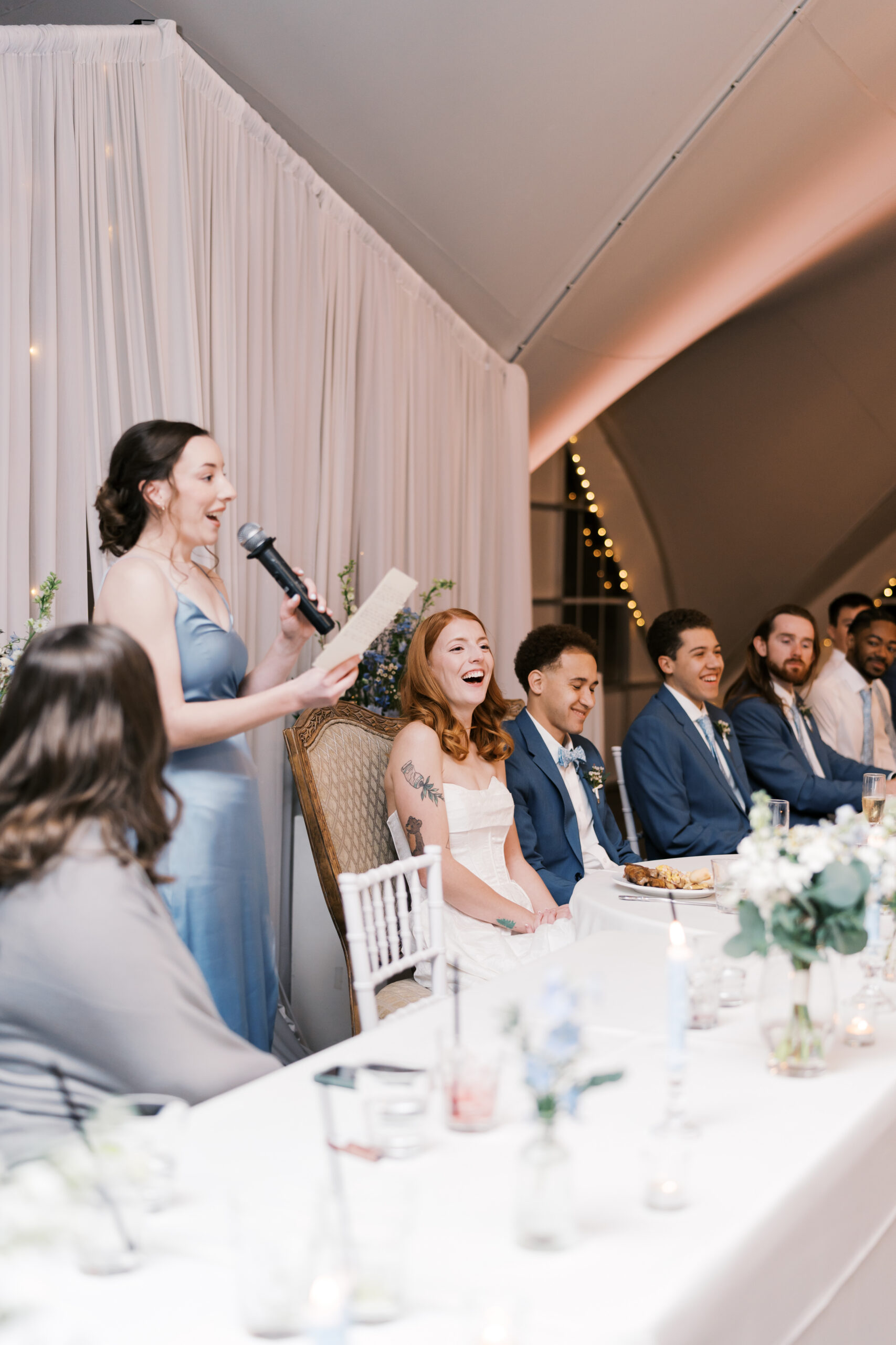 Bridesmaid giving speech with microphone while couple and wedding party sit at head table