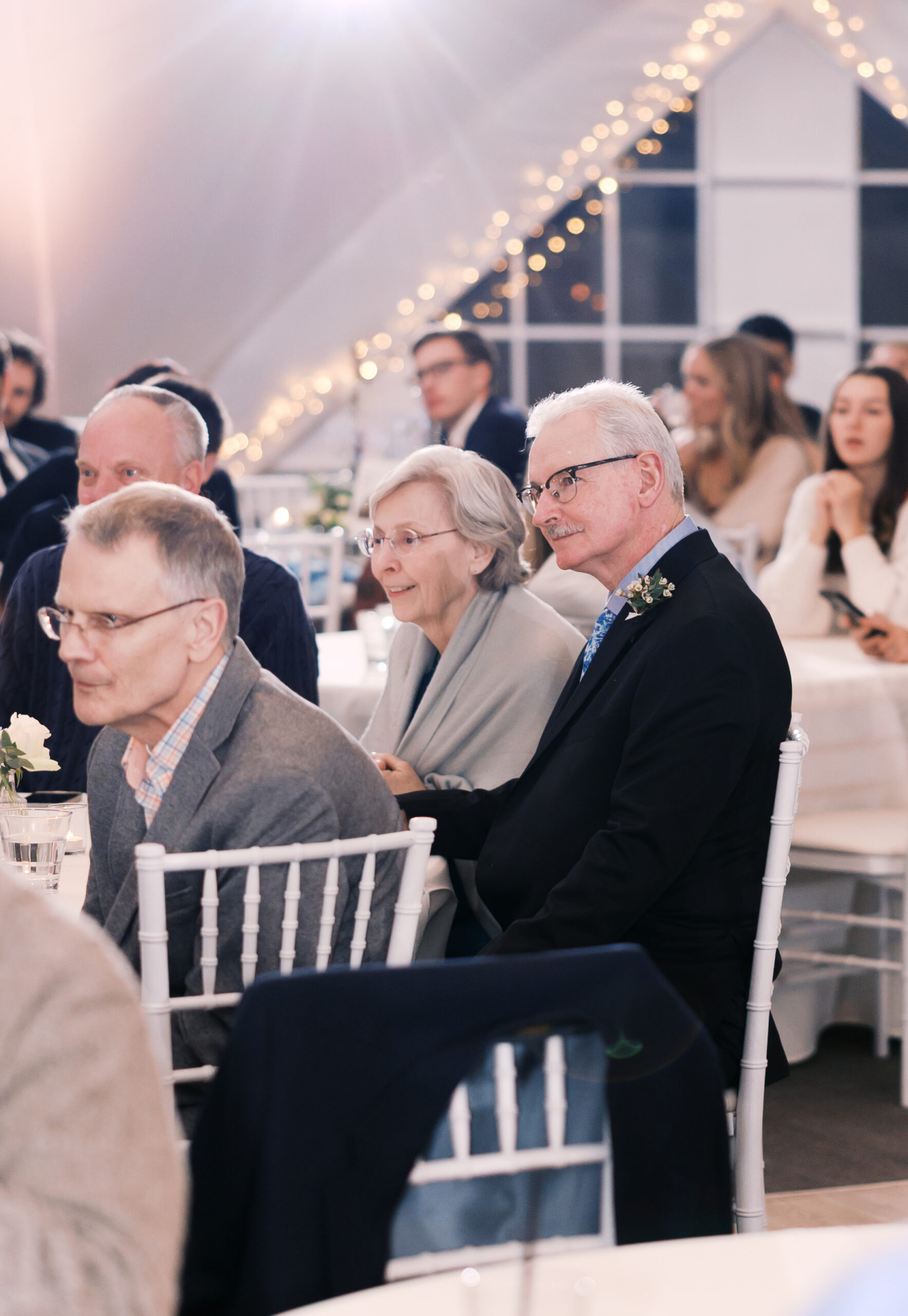 Guests seated at reception tables listening during wedding speeches under warm string lights