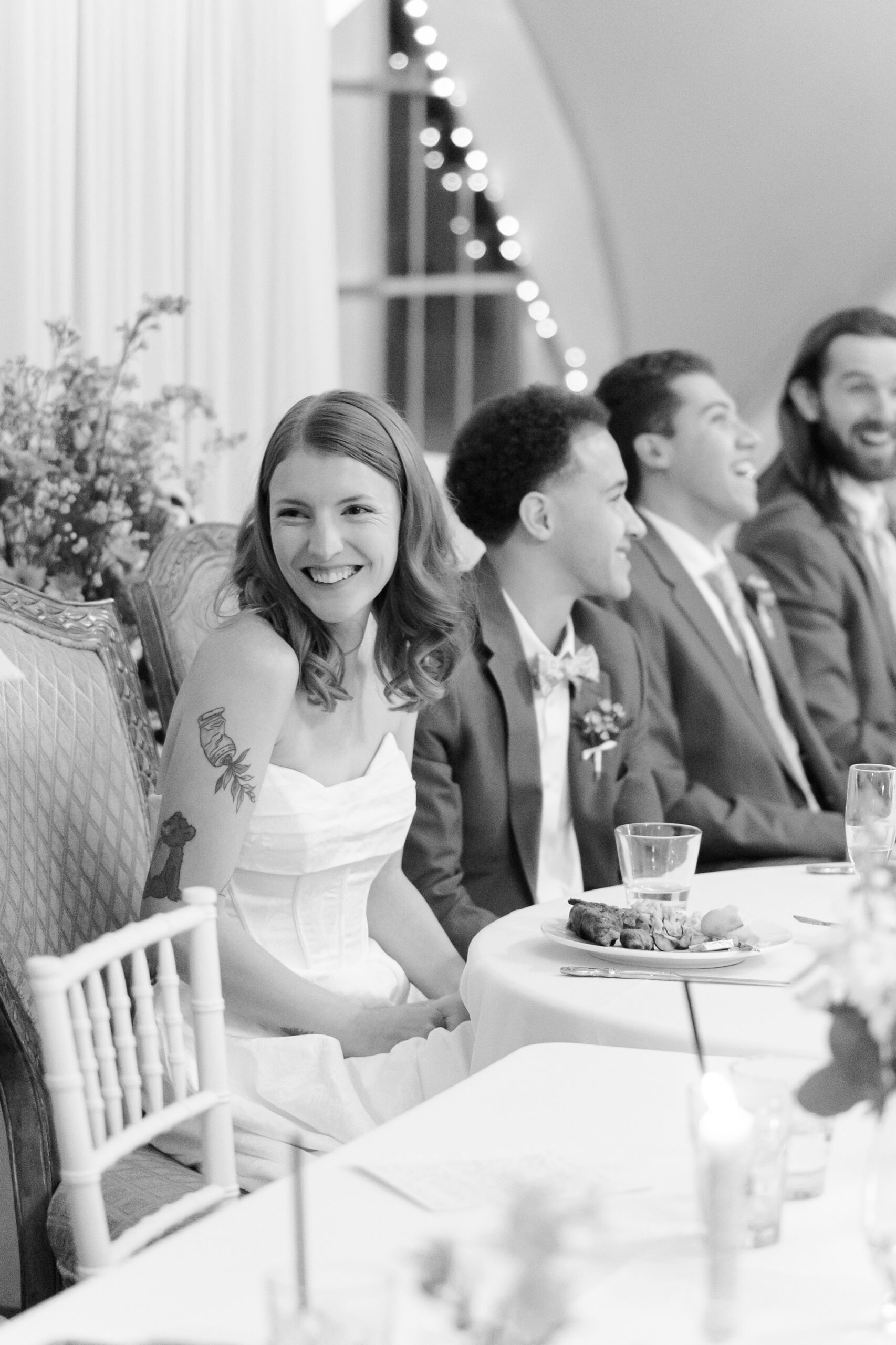 Bride smiling at head table with wedding party during reception, black-and-white photo