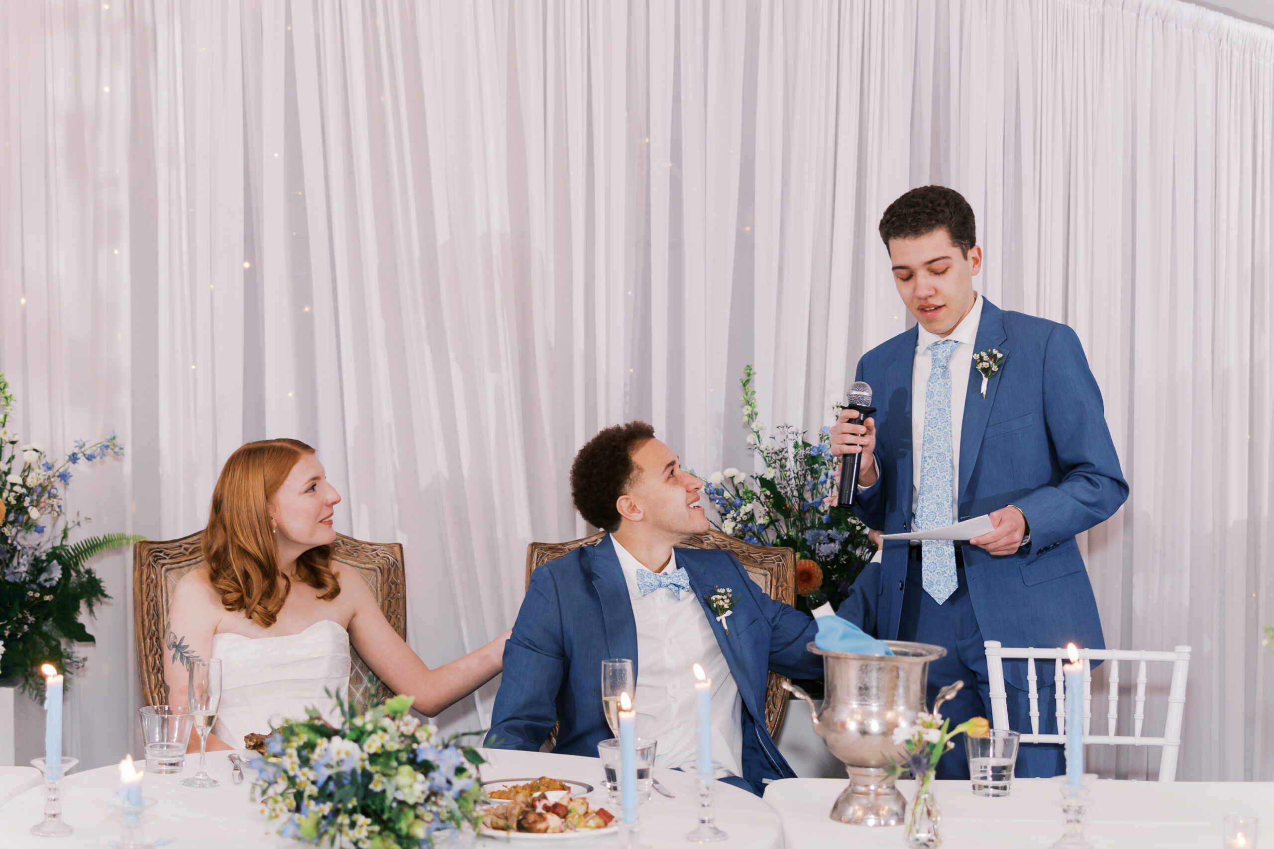 Groomsman in blue suit gives speech at wedding reception while couple sits at decorated head table with candles and flowers.