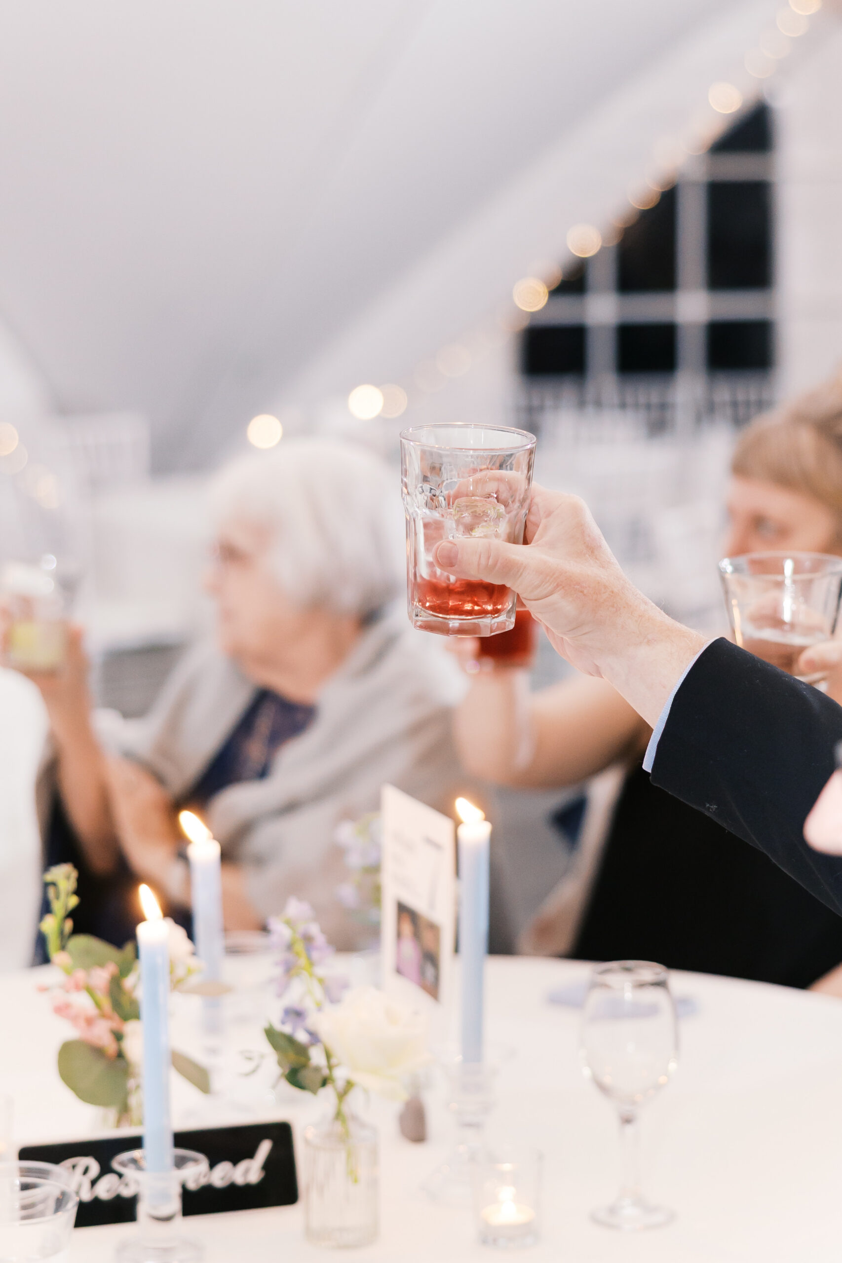 Guest raises glass for toast at wedding reception table with candles, floral decor, and softly lit background.