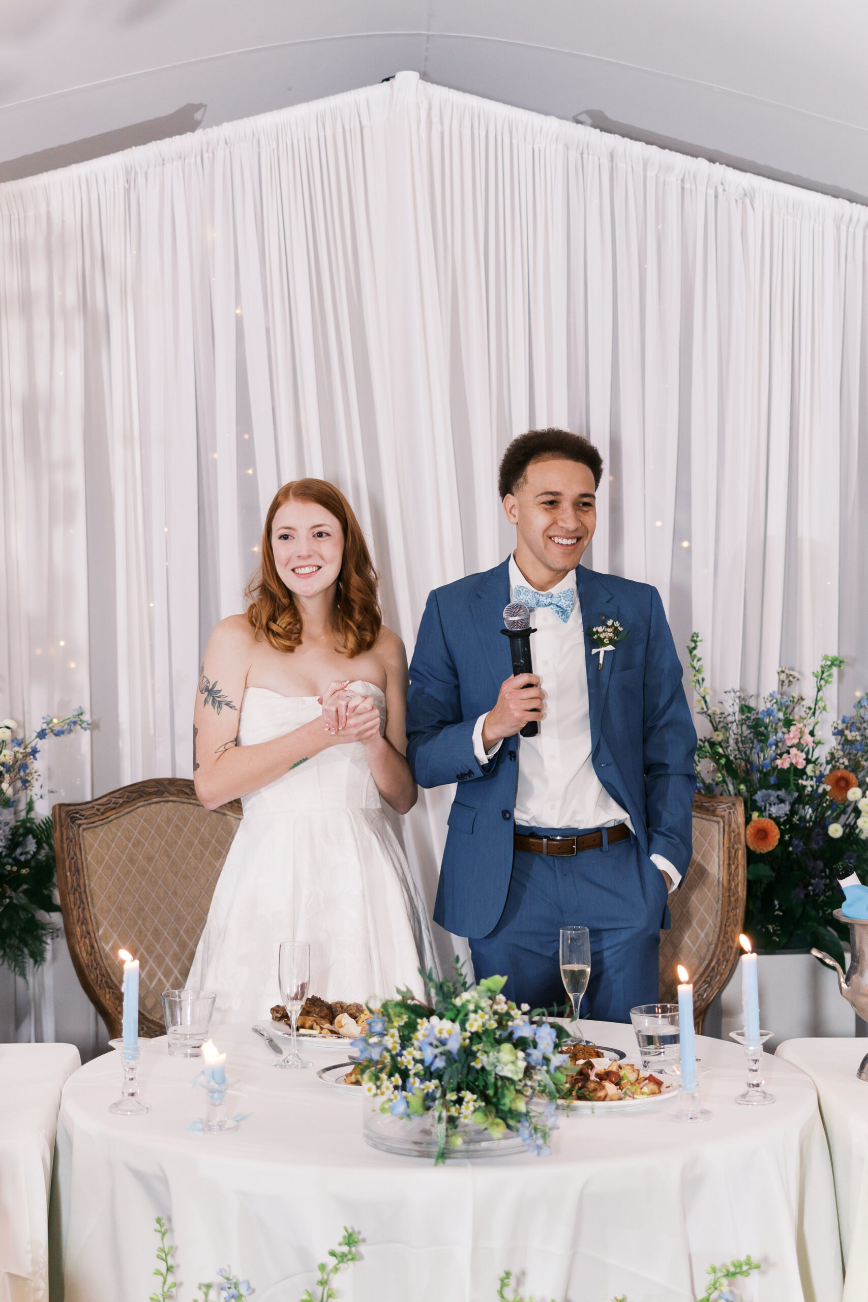 Bride and groom stand smiling at reception head table with candles, florals, and white draped backdrop.