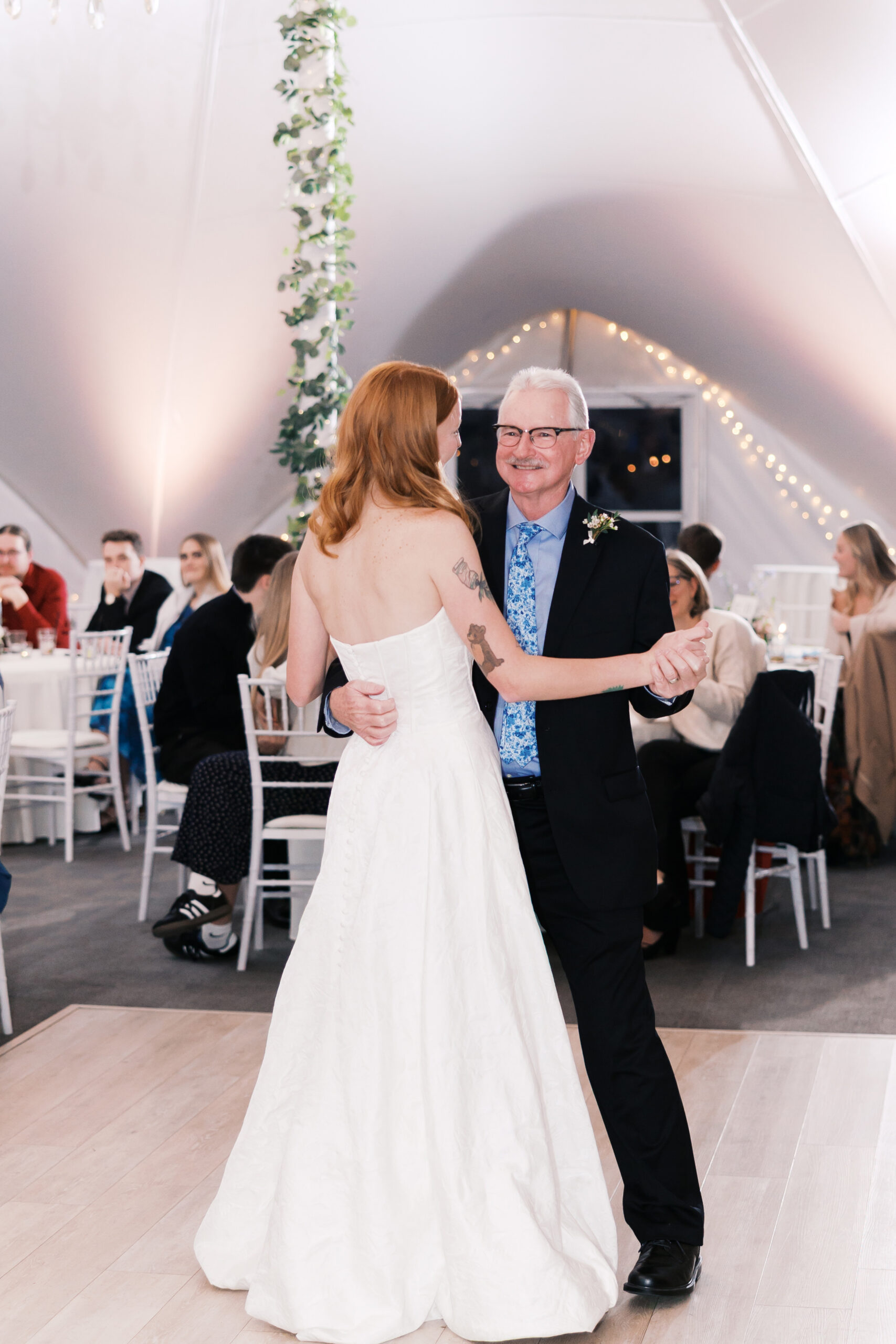 Bride dances with older man on reception dance floor under string lights and guests seated nearby.