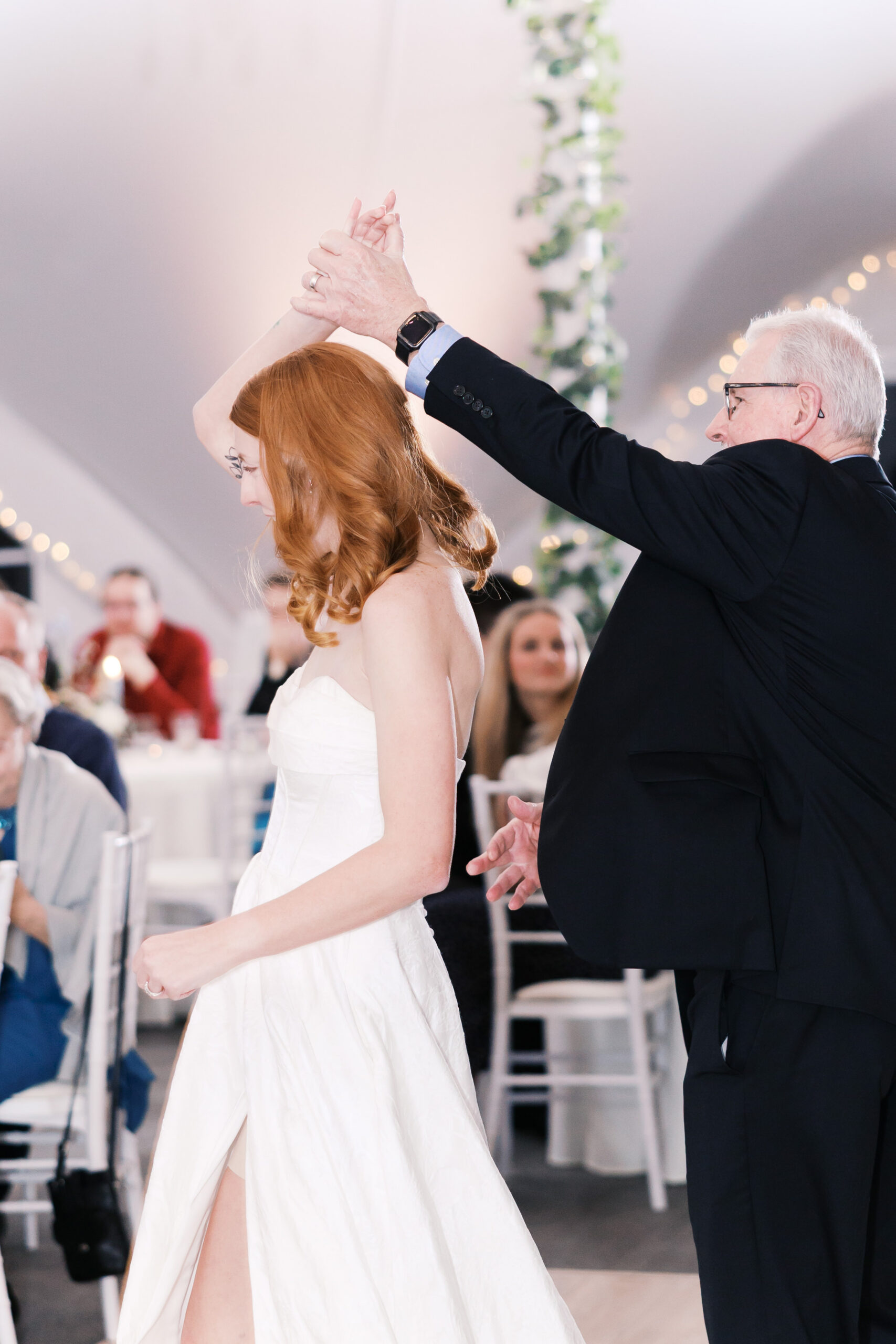 Bride twirls during dance with older man on reception dance floor with guests watching in background.