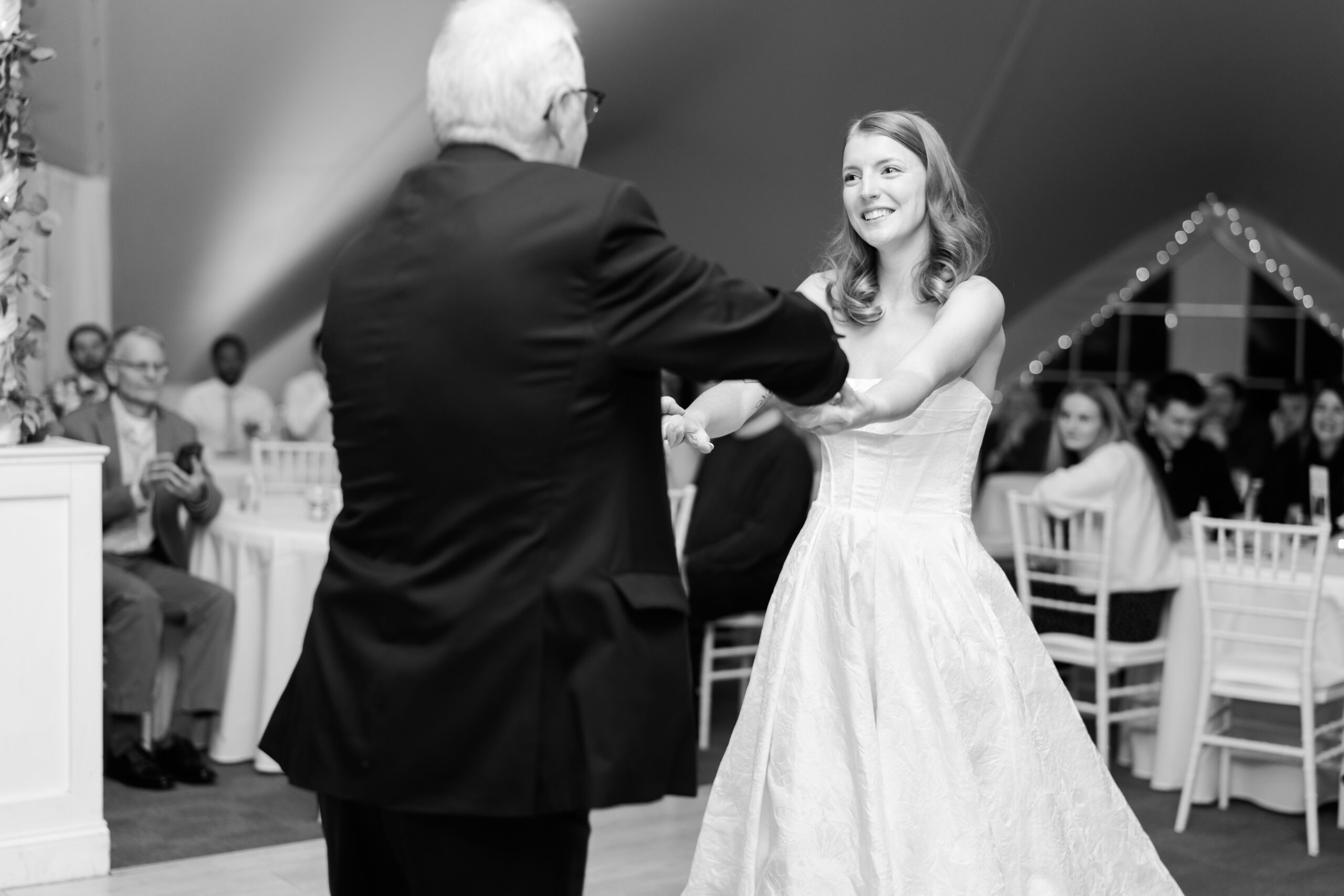 Bride and older man hold hands while dancing at wedding reception with seated guests and soft lighting.